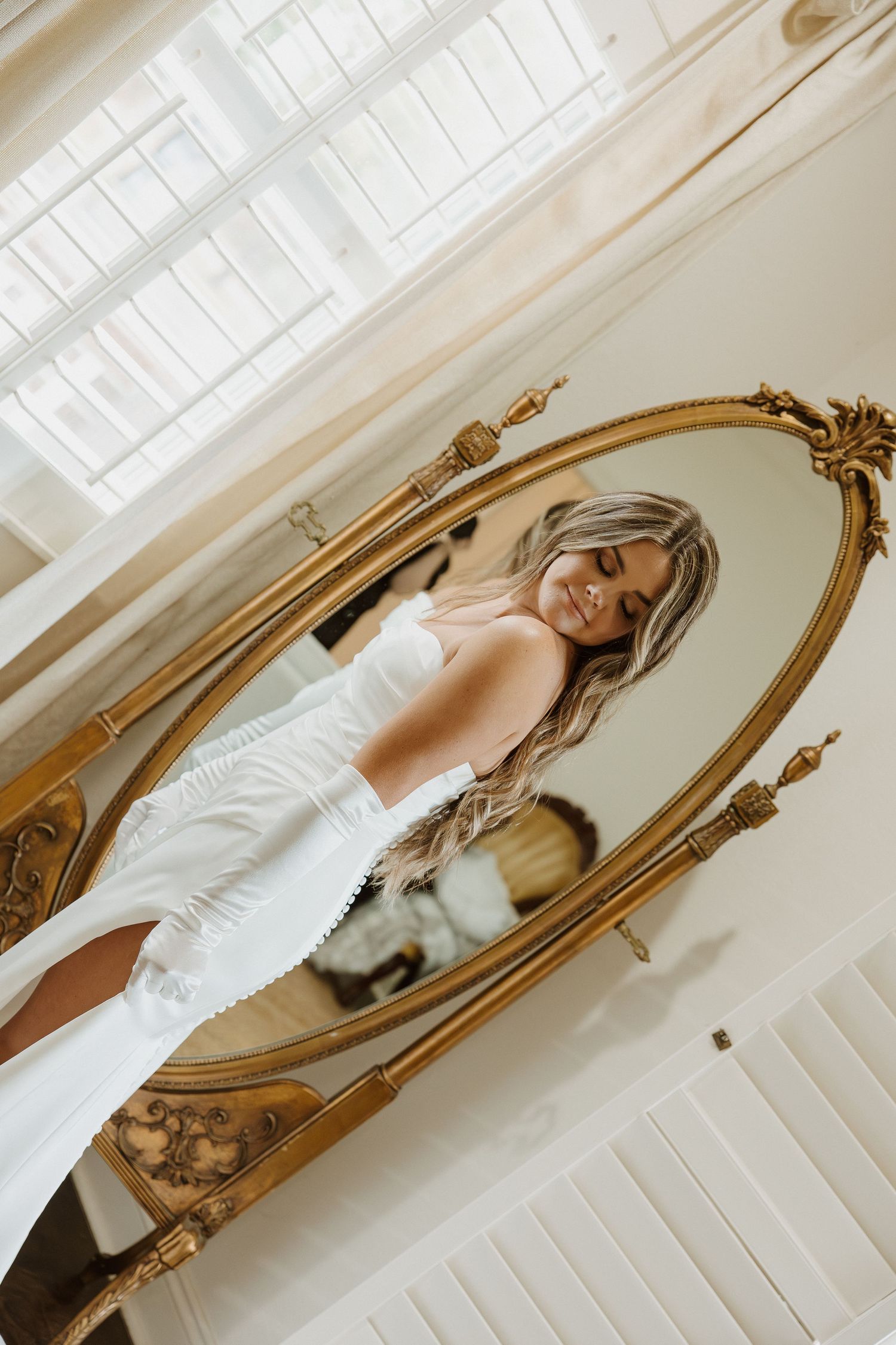 Smiling blonde bride in white dress reflected in ornate gold oval mirror in bright white room.
