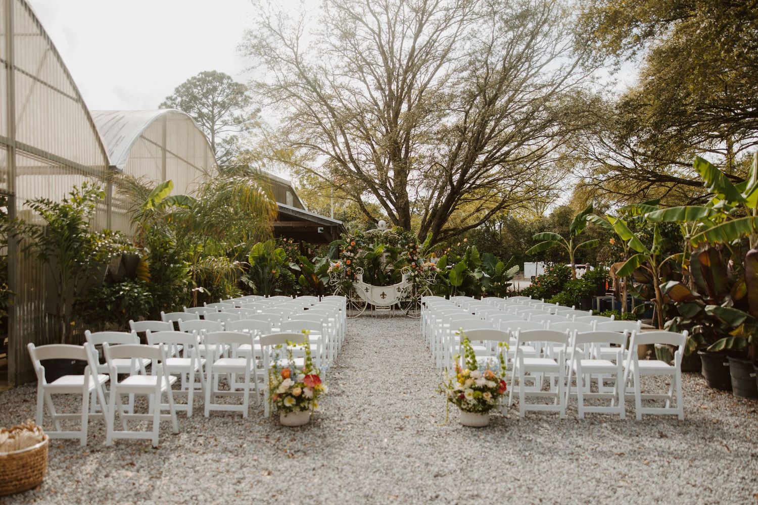 Outdoor wedding ceremony setup with white folding chairs on gravel, surrounded by lush greenery and floral arrangements.
