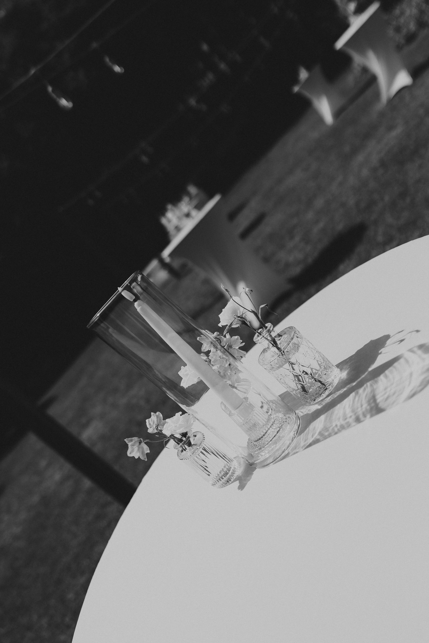 Black and white photo of elegant glass vase centerpiece with flowers on a white table at an outdoor event.