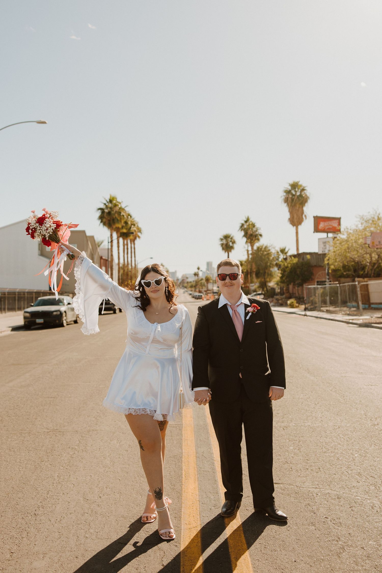 A couple stands in the middle of a palm tree-lined street during a sunny wedding photoshoot.