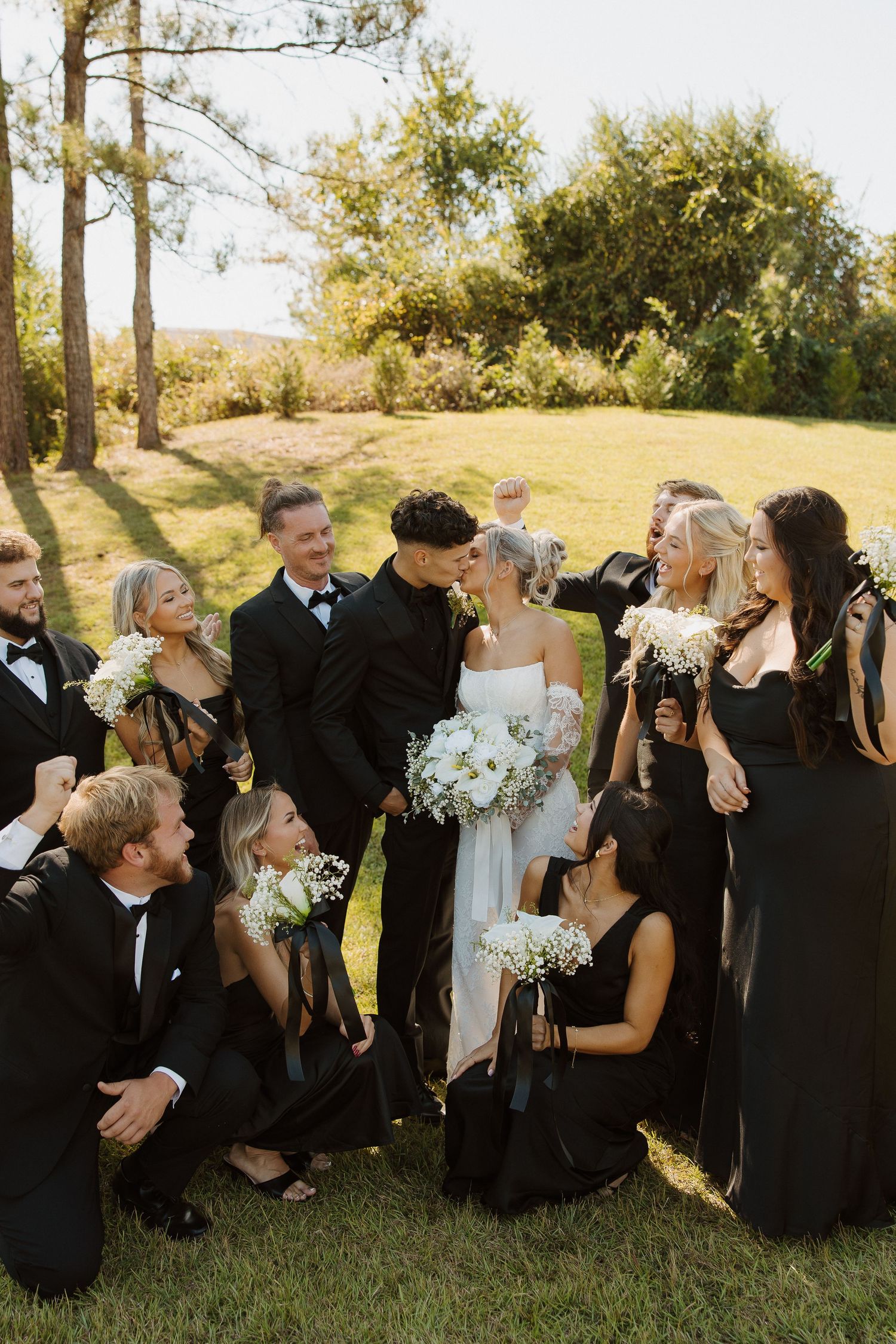 Wedding party in black and white attire gathered for a group photo on a sunny lawn.