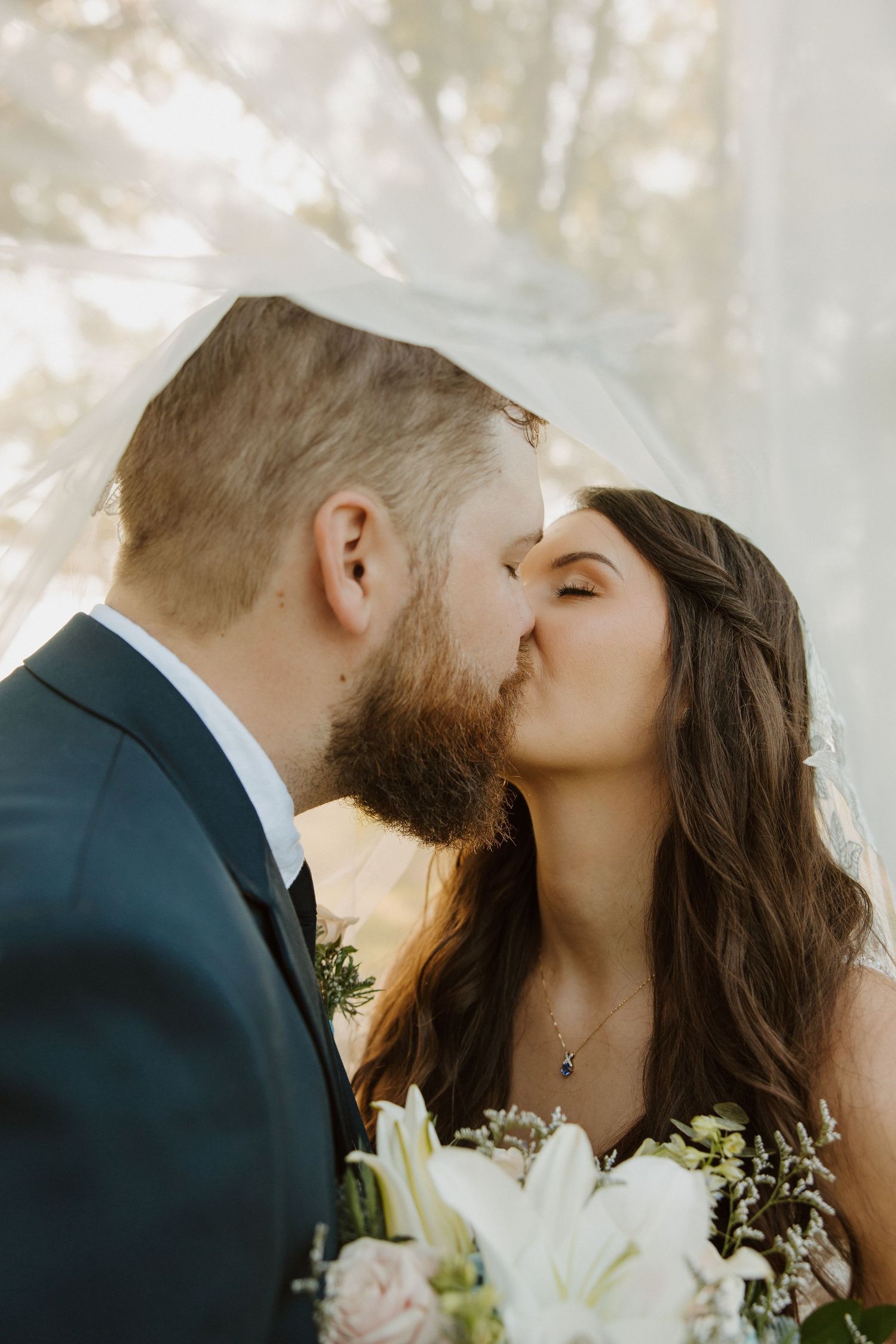 A bride and groom share a romantic kiss under a veil, with the bride holding a white floral bouquet on their wedding day.