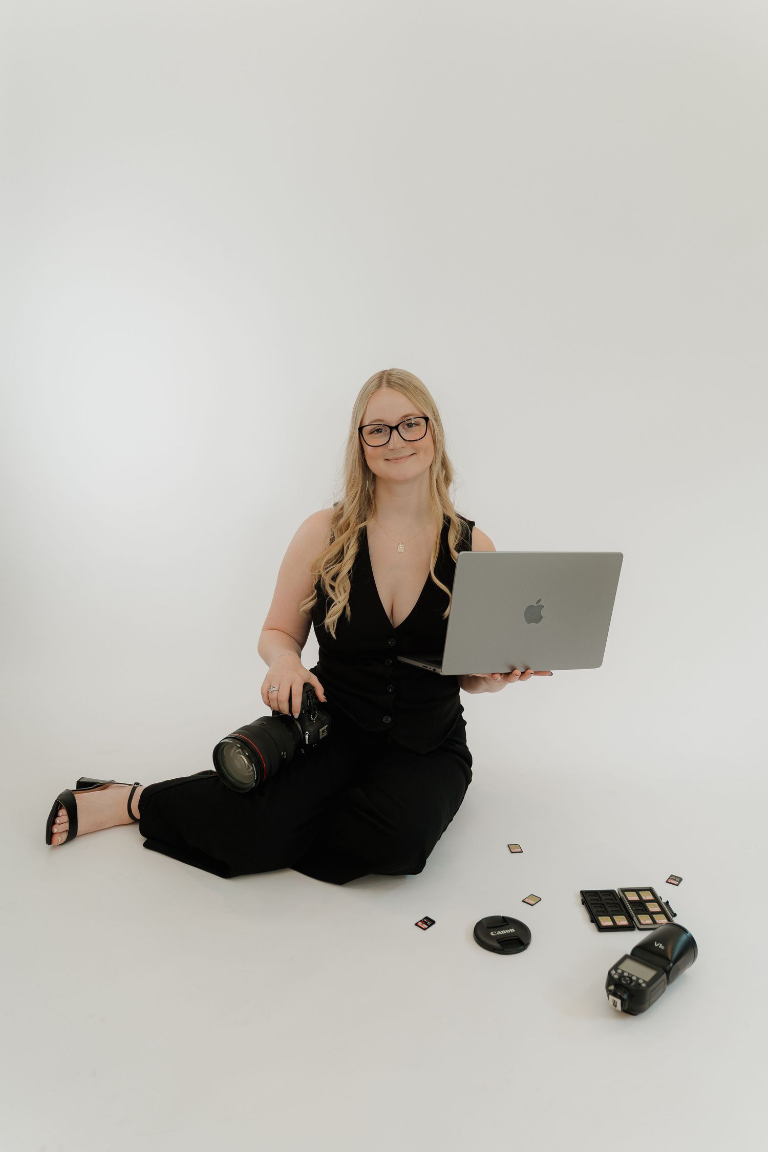 Professional photographer in black outfit sits on white floor with laptop and camera equipment during studio photoshoot.