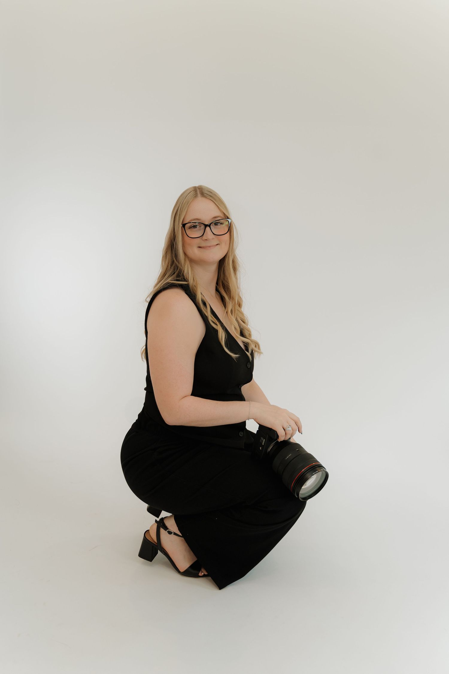 Professional photographer in a black dress kneels with camera against white studio backdrop.