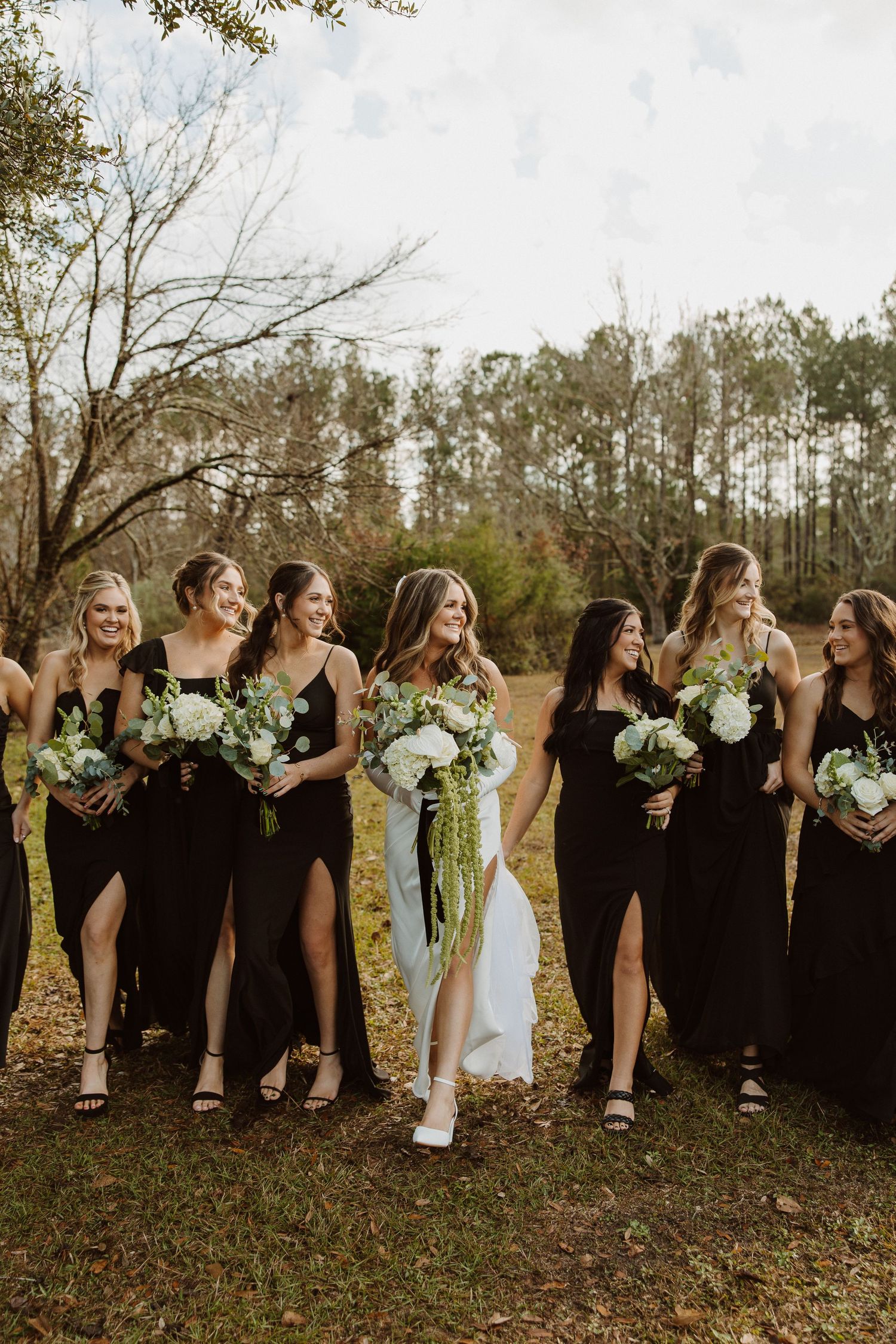 A bridal party in black dresses and one white gown pose together outdoors in a candid laughing moment.