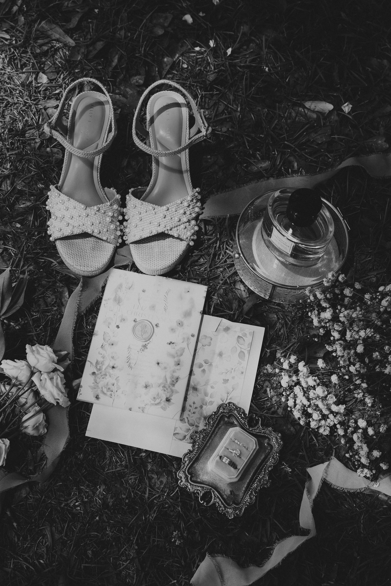 Black and white flat lay of bridal accessories including pearl sandals, wedding invitation, rings, and baby's breath flowers.
