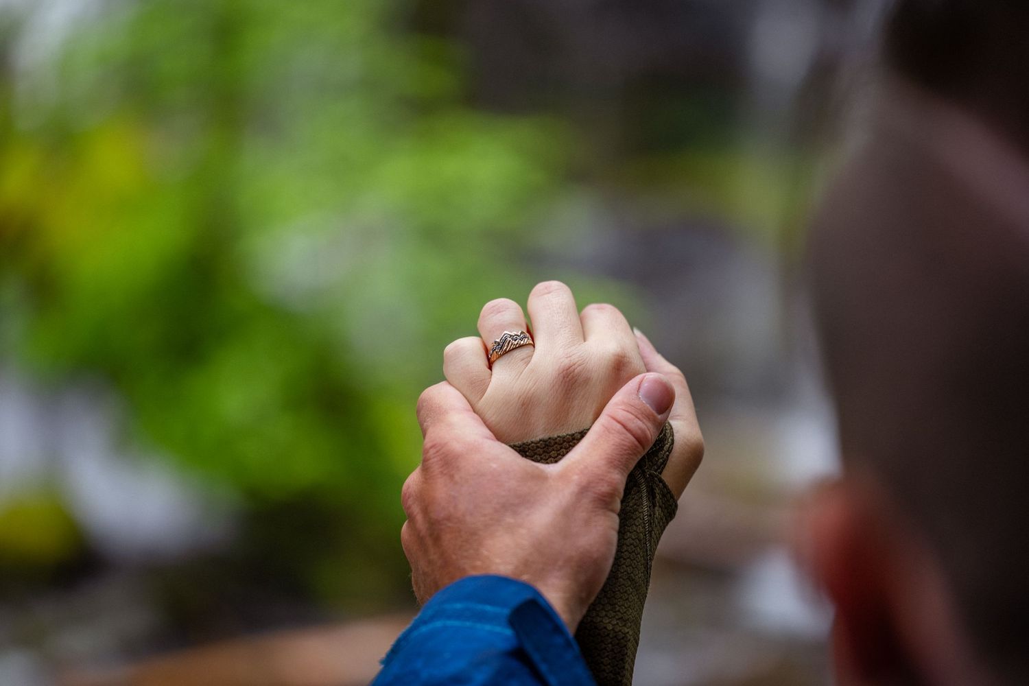 Close-up of two hands intertwined, one wearing a diamond engagement ring, with lush green nature in the background.
