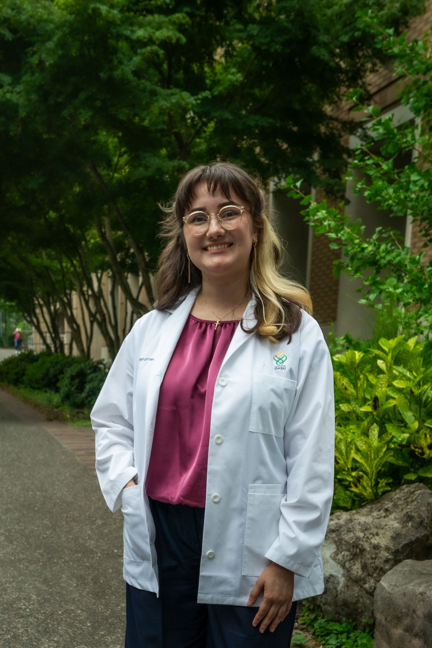 Medical professional wearing burgundy top and white coat photographed in a garden setting.
