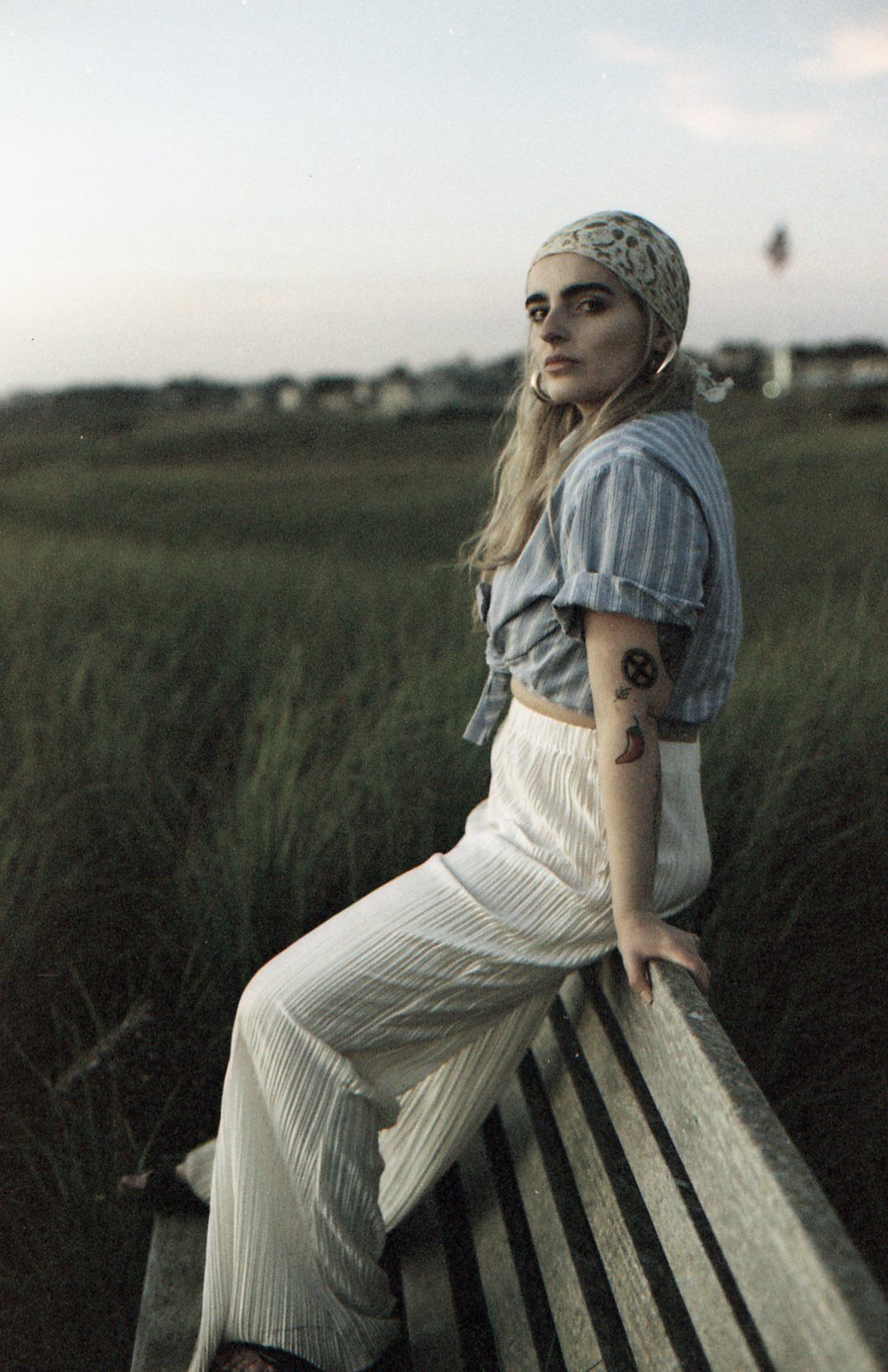 A person in a white dress and gray headscarf sits contemplatively on a wooden bench in a field at dusk.