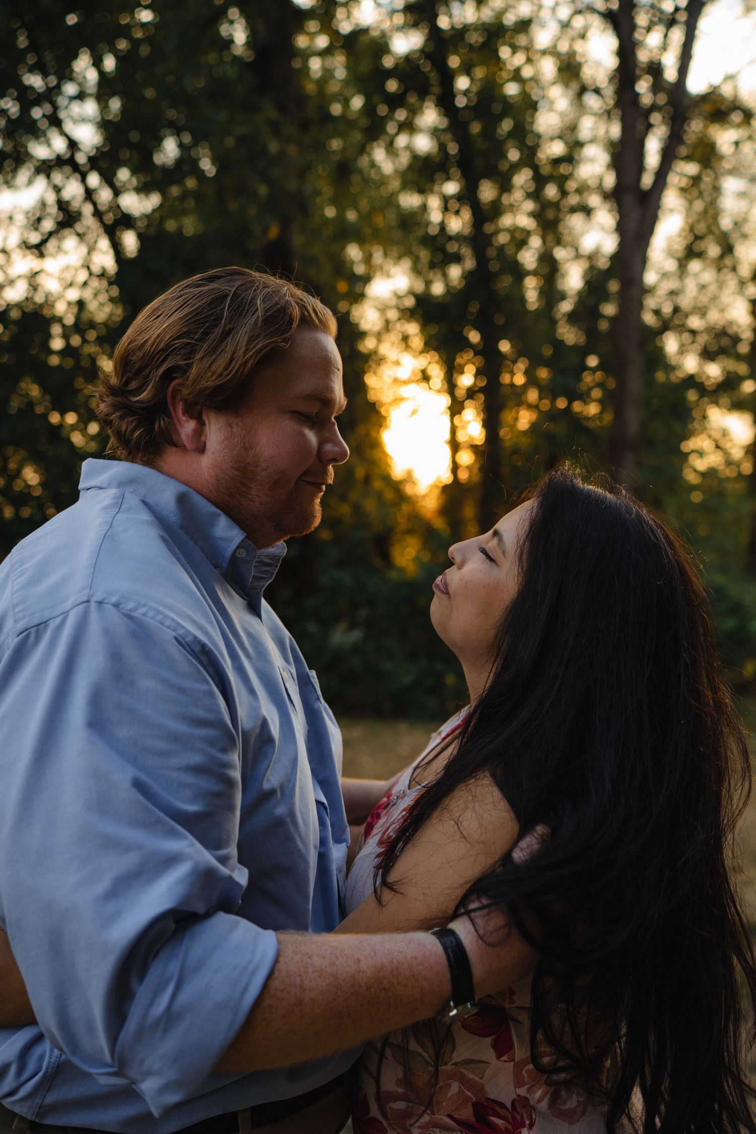 A couple shares an intimate moment at sunset under trees decorated with lanterns.