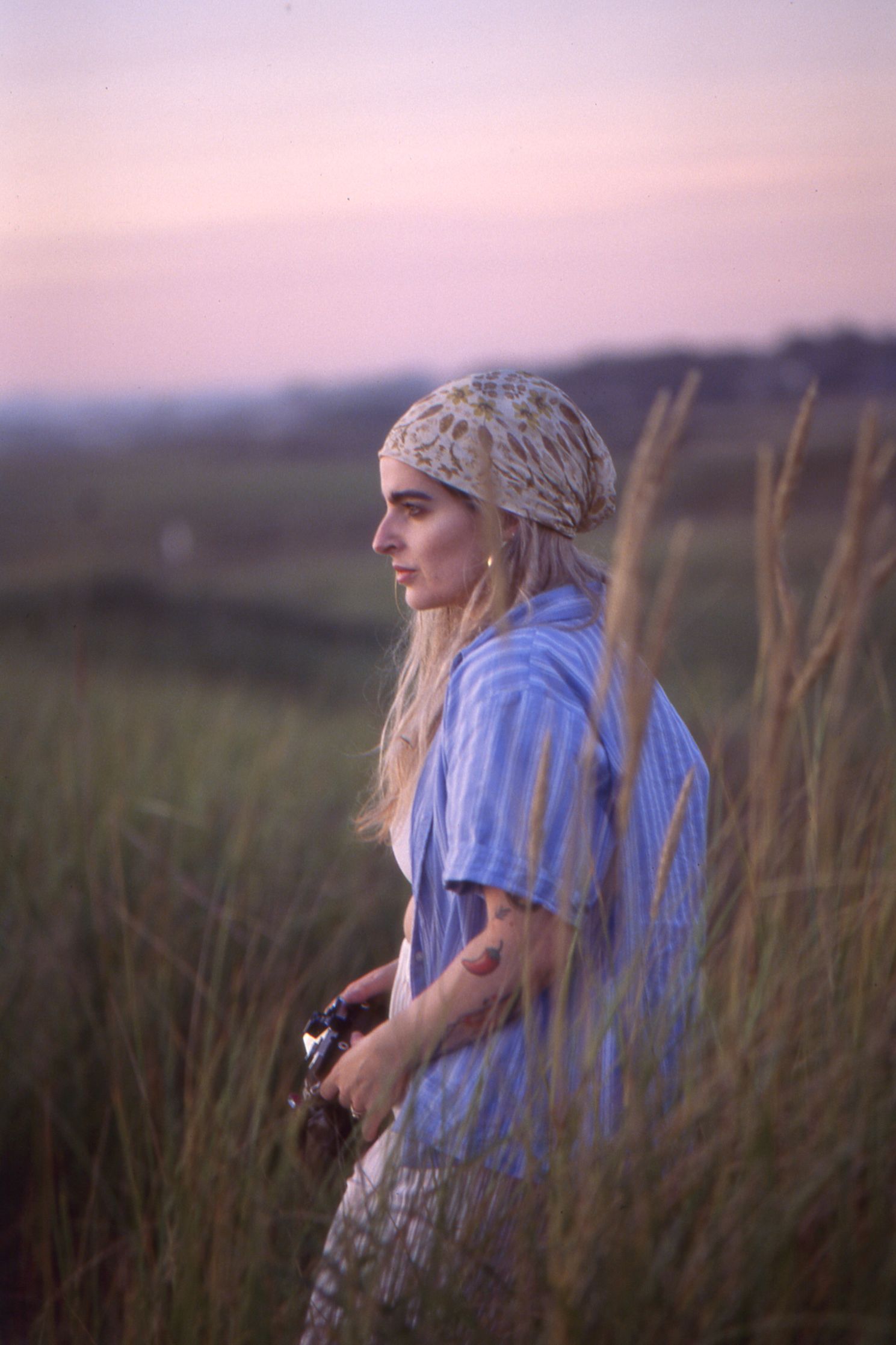 A person wearing a blue shirt and bandana sits in tall grass during sunset with a soft pink sky in the background.