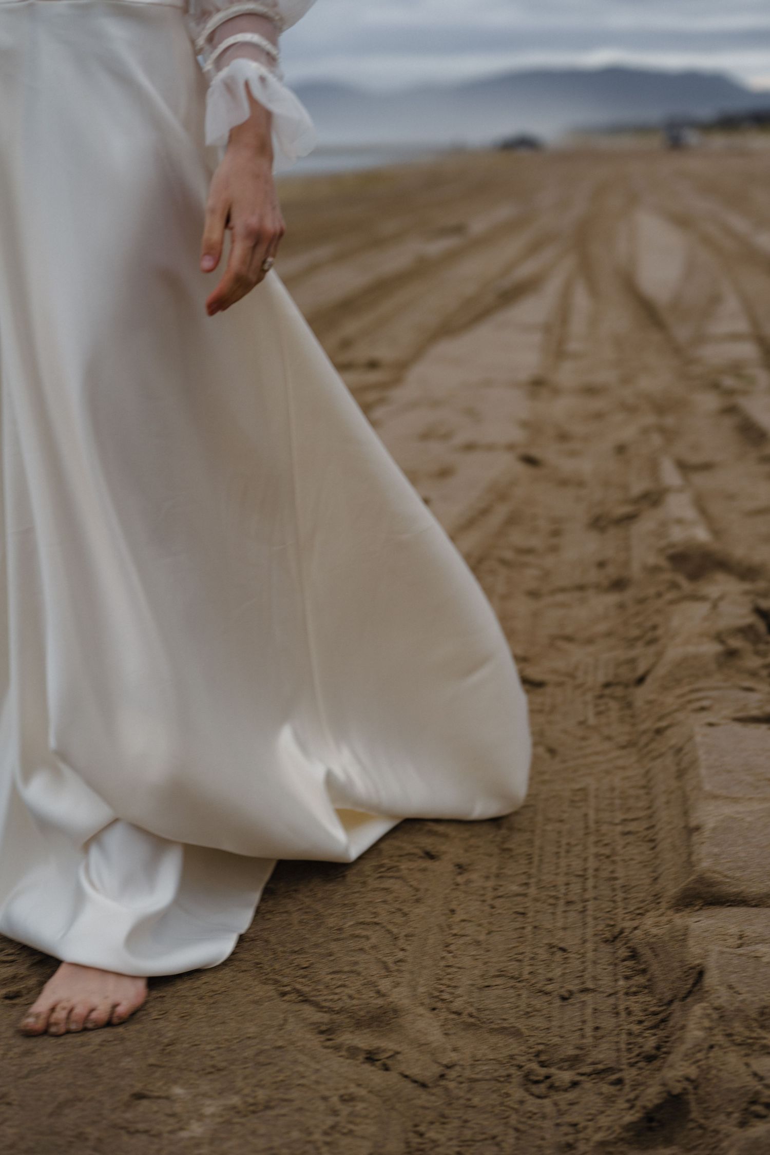 Close-up view of bare feet walking on sandy beach in flowing white wedding dress hem.