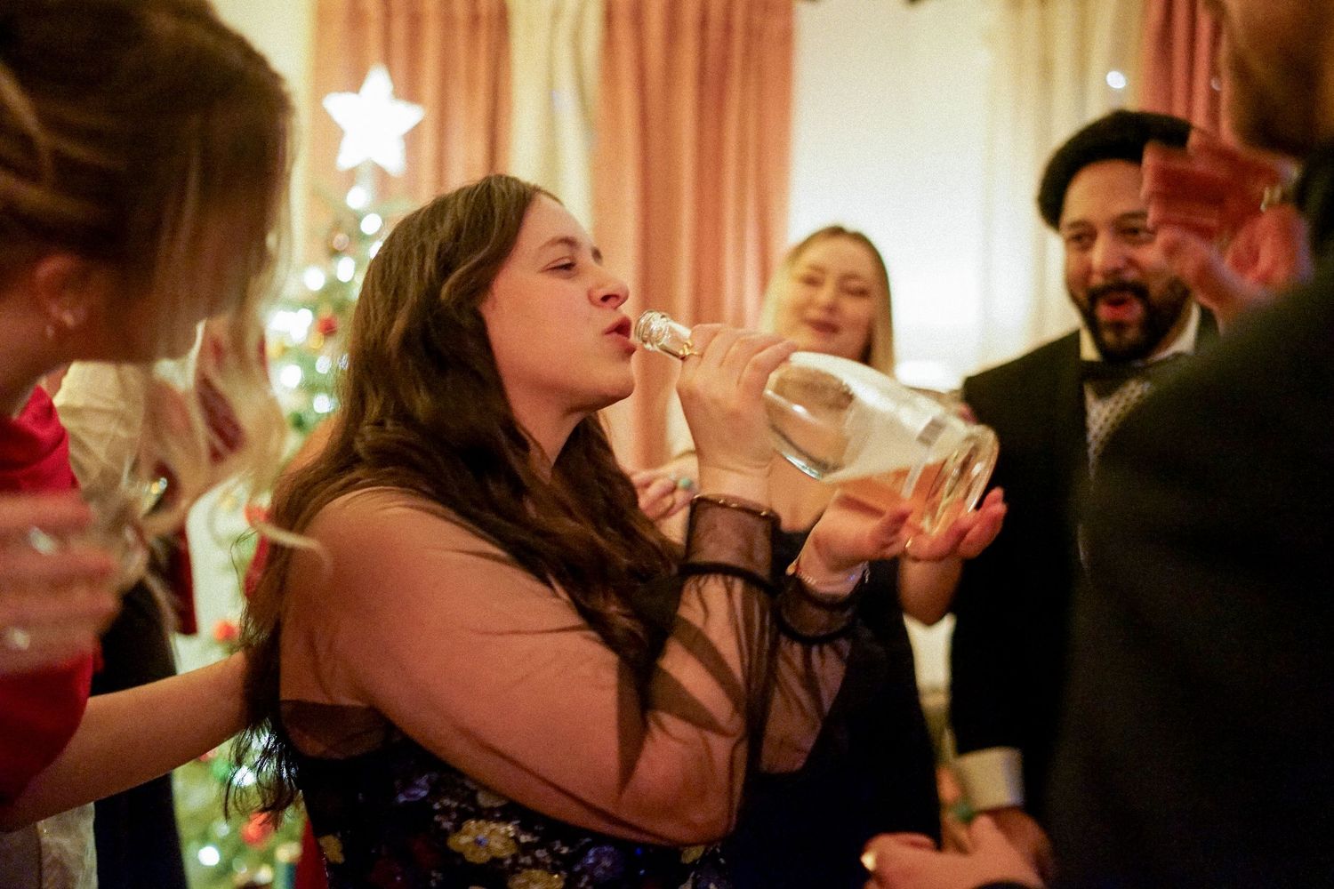 Party guests drink from bottles at a festive indoor celebration with string lights.