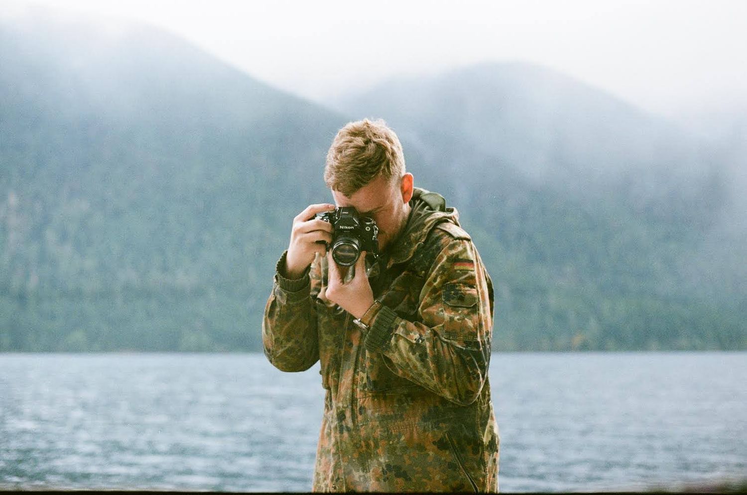 A photographer in camouflage clothing takes photos by a misty mountain lake.
