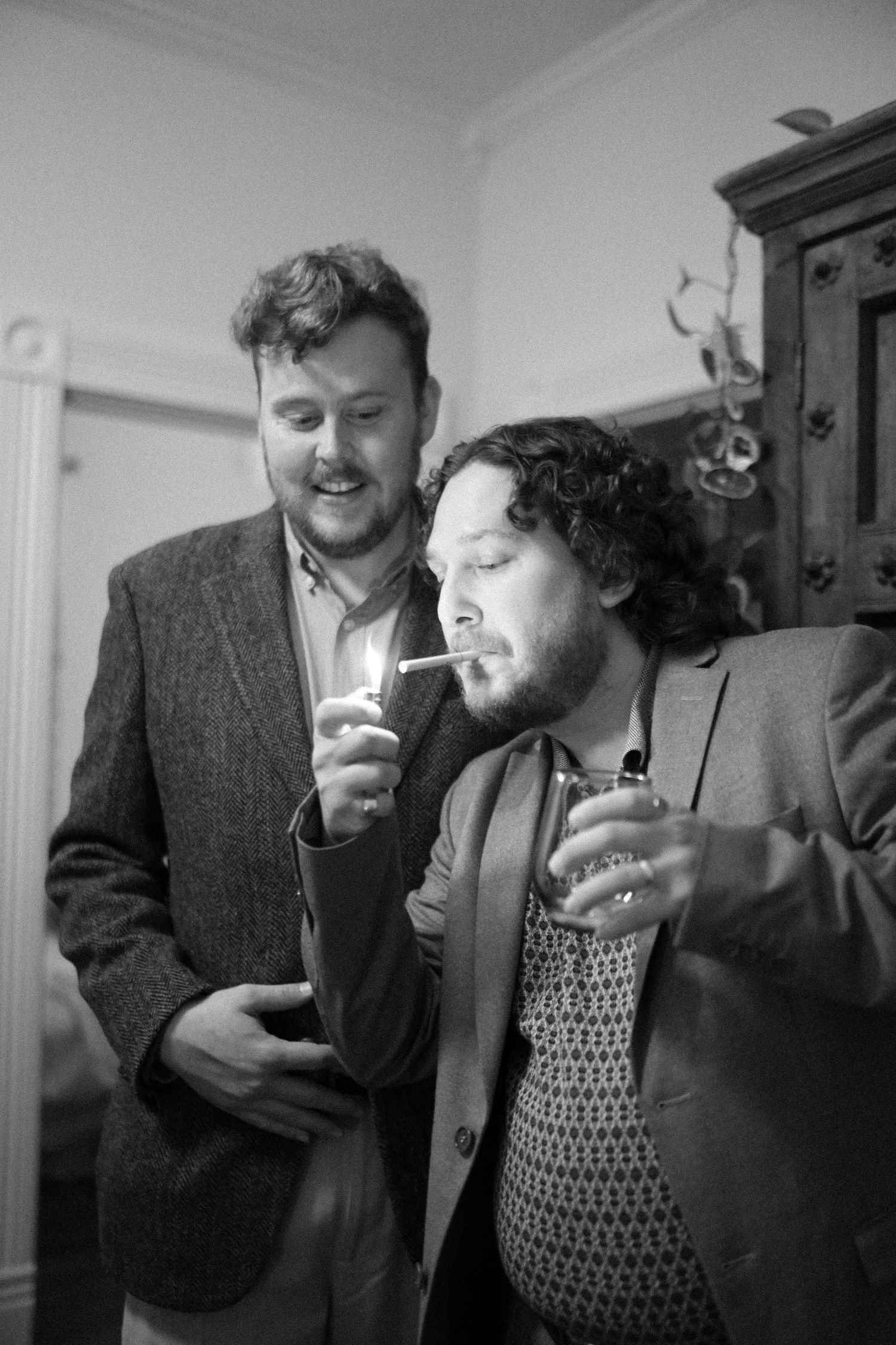 Two friends in suits sharing drinks and laughter at indoor gathering.