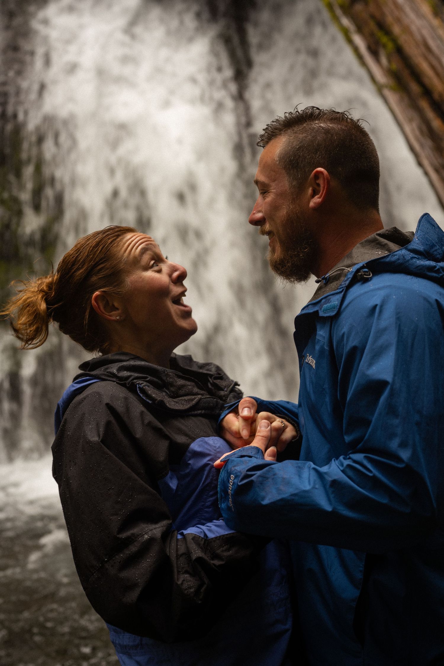 Couple holding hands and laughing together in front of a waterfall, wearing blue and black rain jackets.