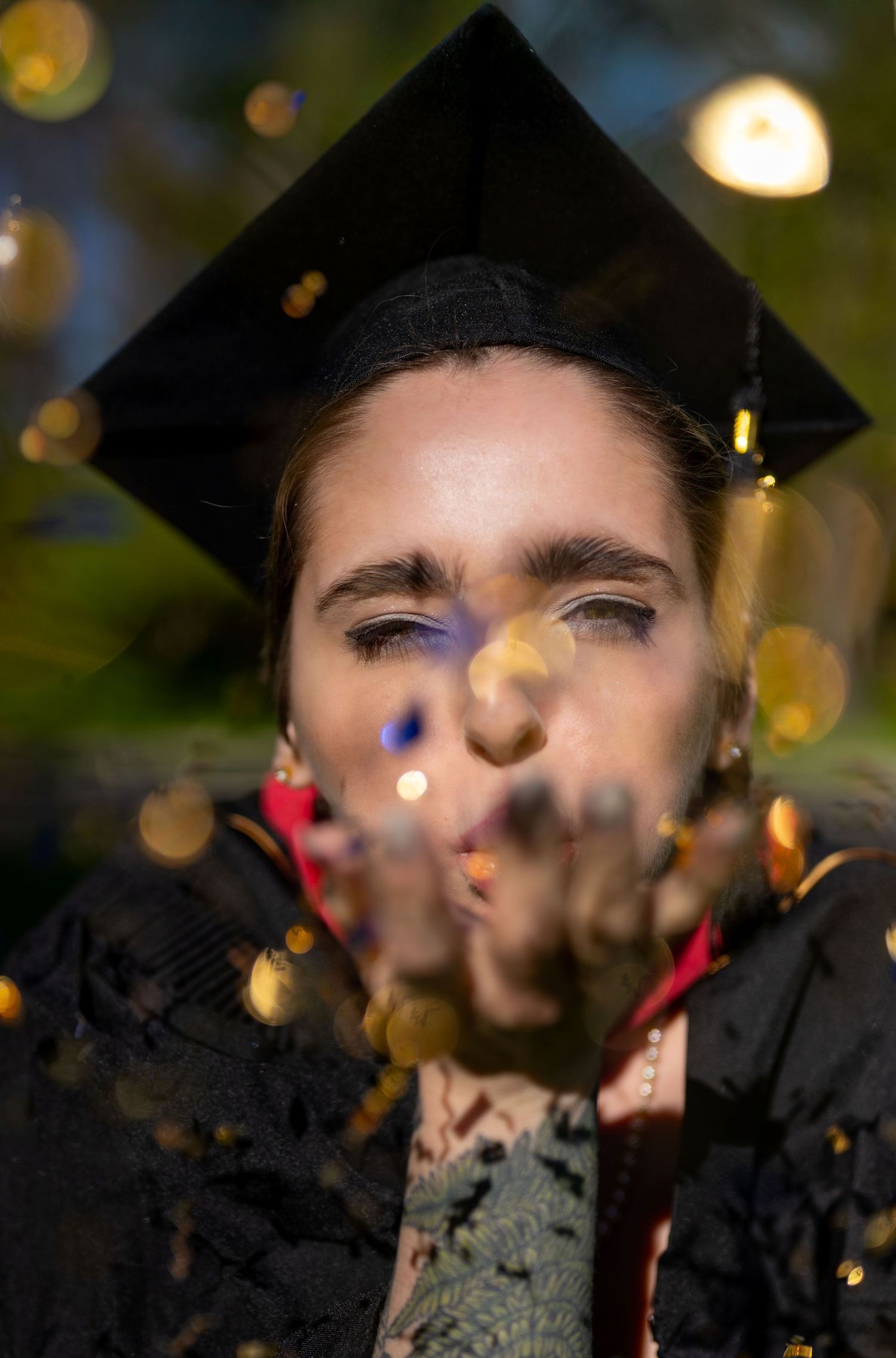 Graduate in cap and gown with confetti falling in dark lighting.