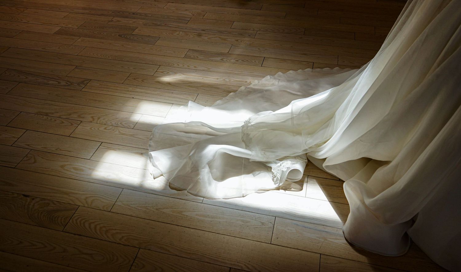 Flowing white wedding dress train on hardwood floor with sunlight streaming through windows.