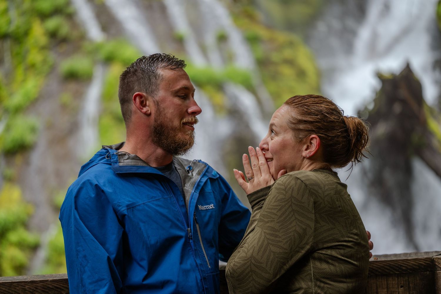 Couple sharing a romantic moment in front of a stunning waterfall, wearing rain jackets in a lush green outdoor setting.