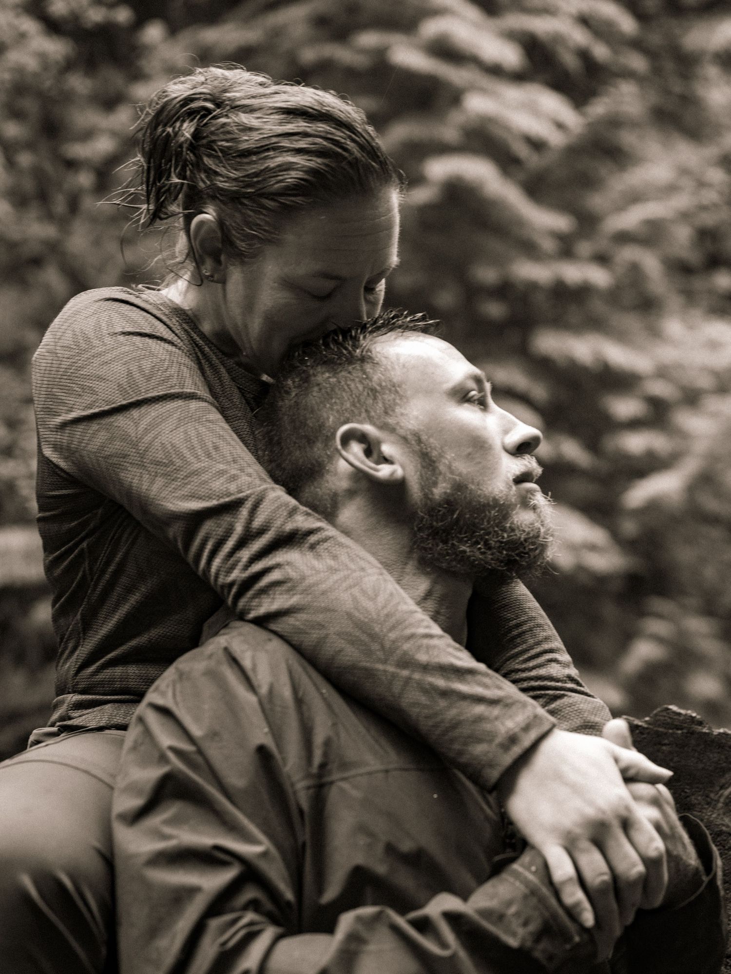 A woman embraces a bearded man from behind in a tender black and white outdoor portrait.