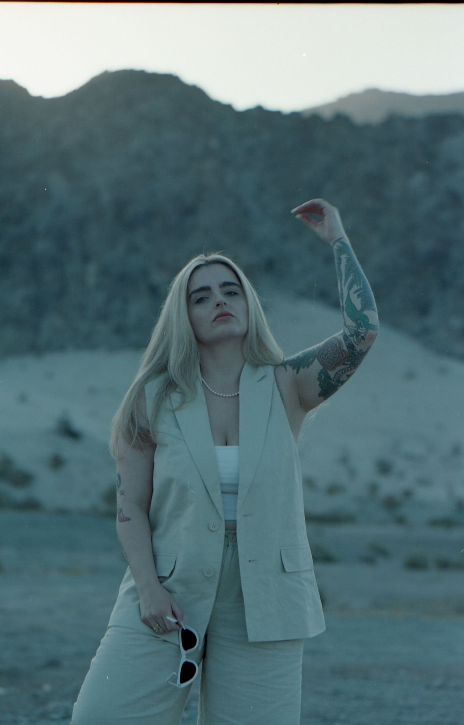 Person in light colored outfit stands in desert landscape with mountains in background.