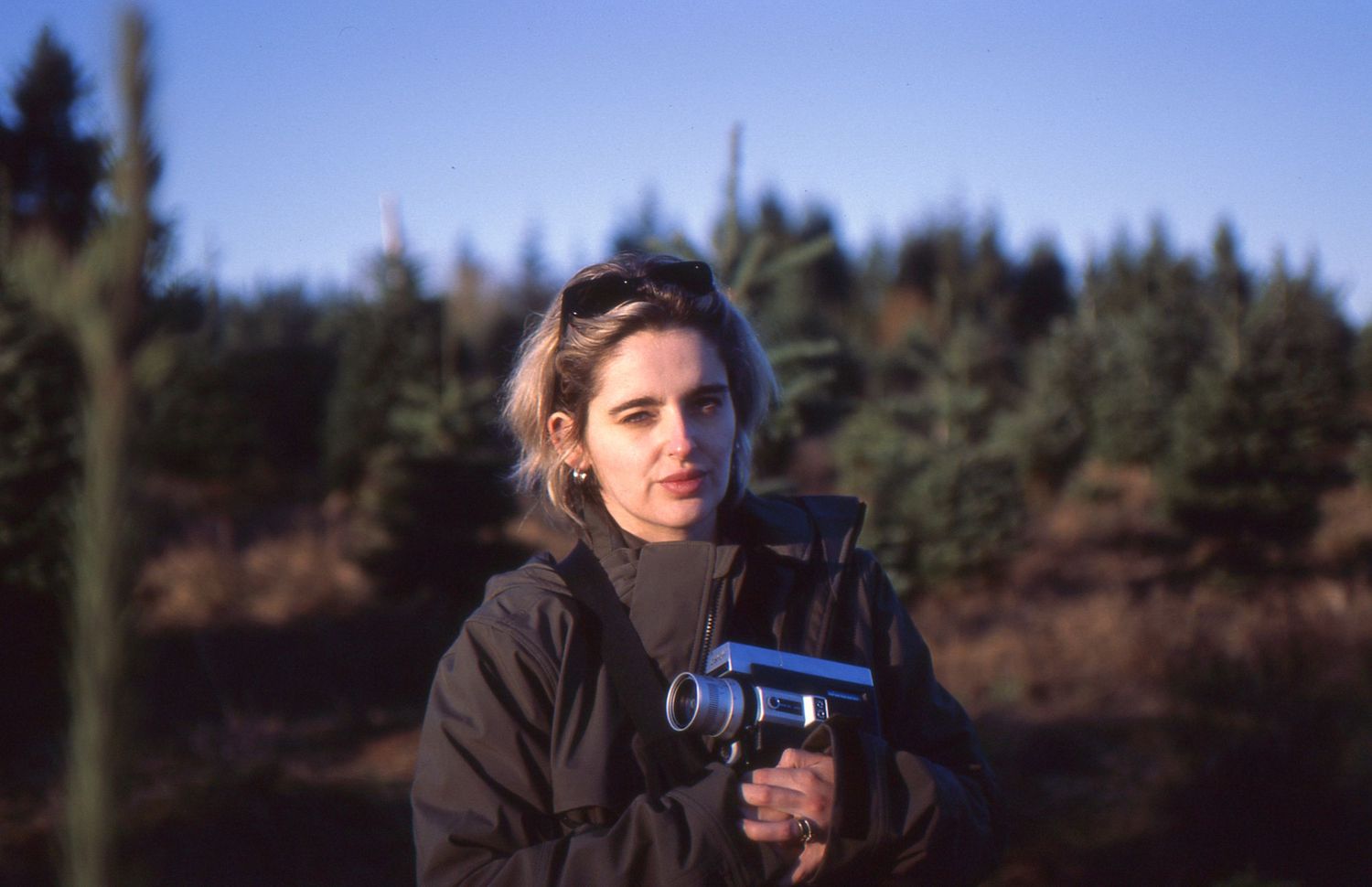 Person in dark clothing walking outdoors in a wooded area on a sunny day with blue sky visible.