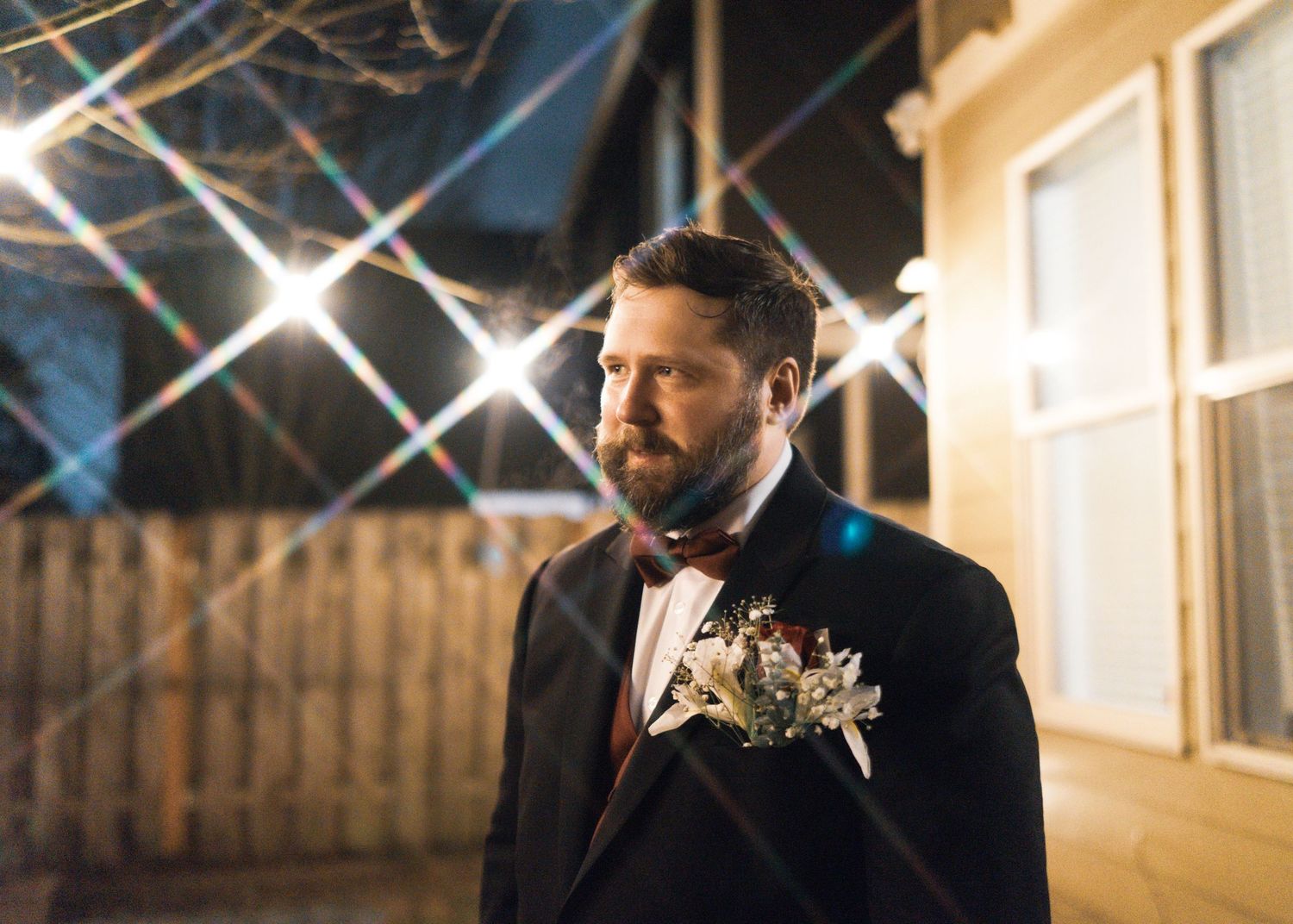 A man in formal attire stands in front of a white building with string lights overhead.