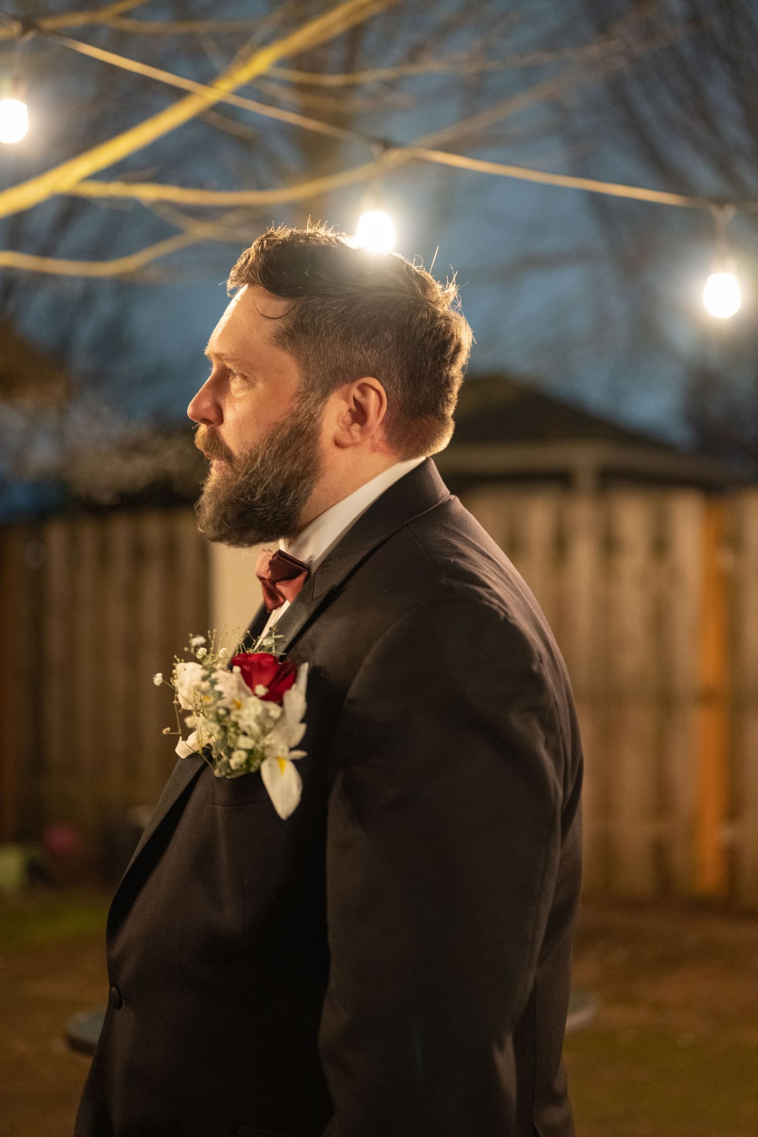 A groom in a black suit with a red boutonniere stands under twinkling lights at an evening wedding ceremony.