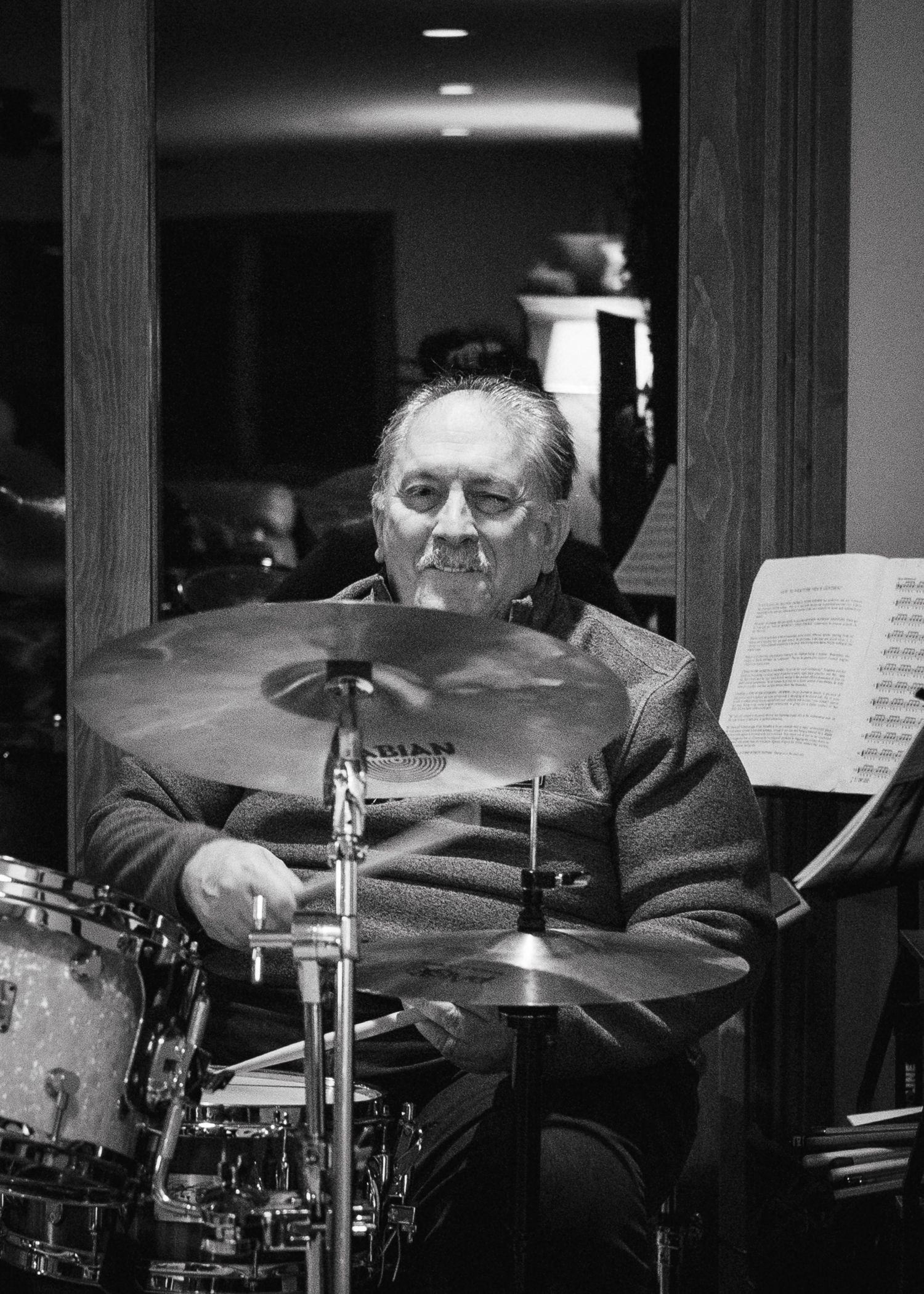 Drummer playing an acoustic drum set with sheet music stands visible in the performance space.