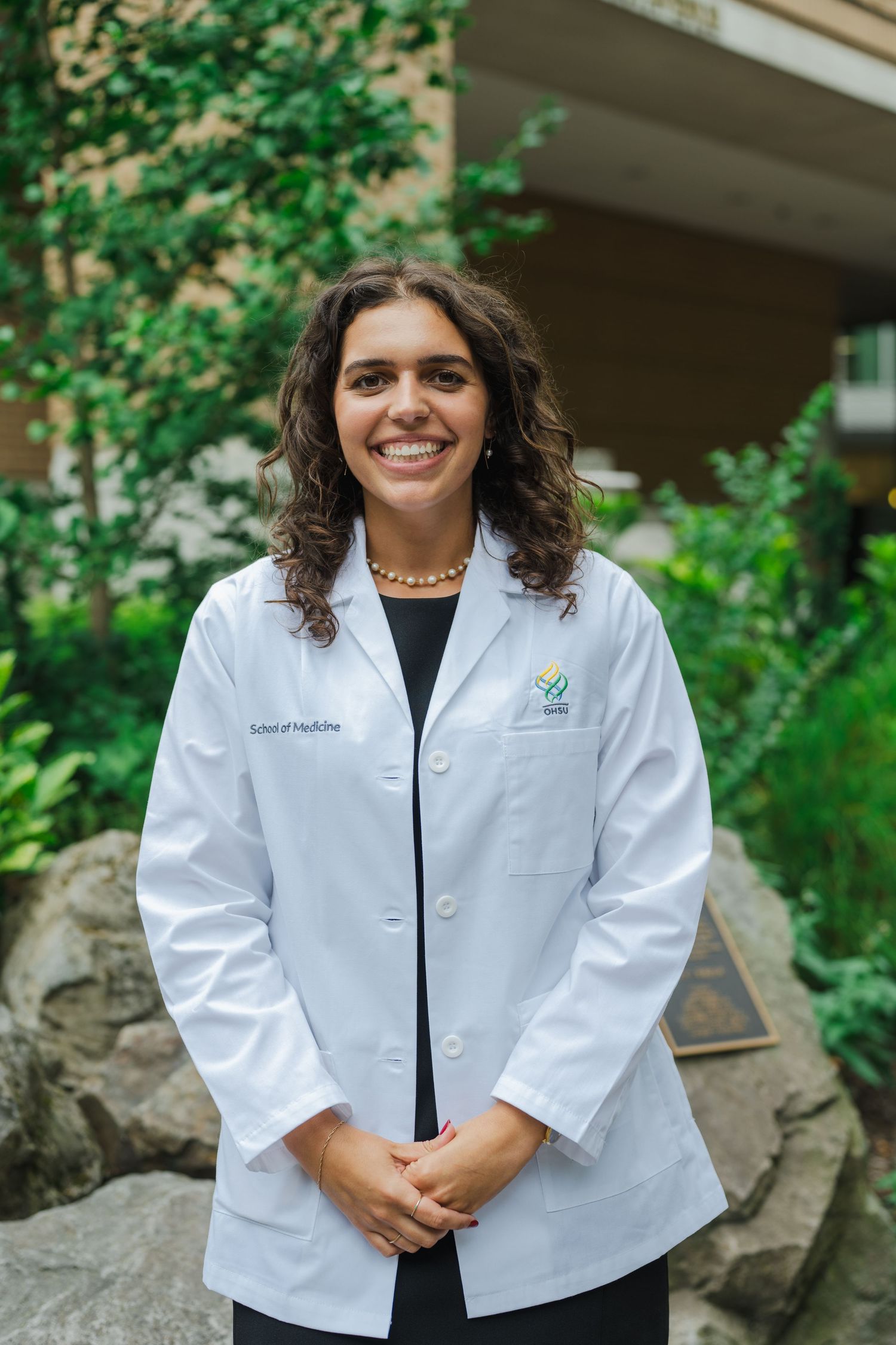 Healthcare professional in a white lab coat smiling warmly against a natural outdoor backdrop.