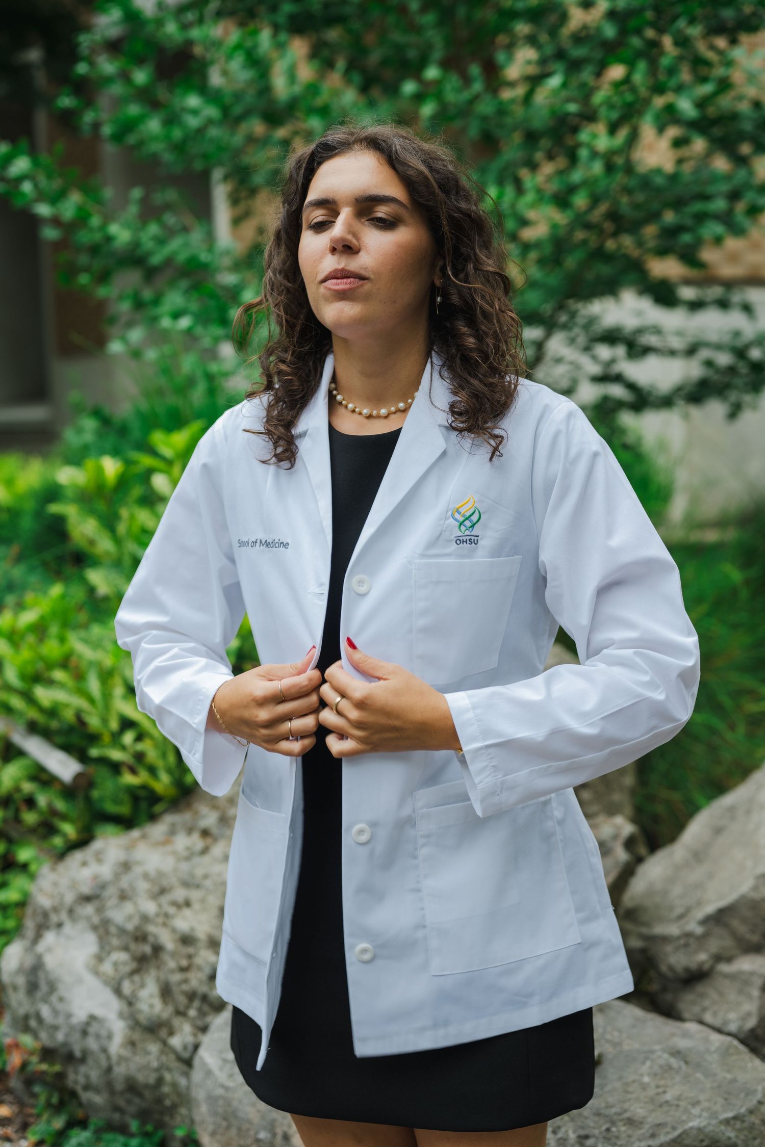 Person wearing a white lab coat standing outdoors against greenery and rocks.