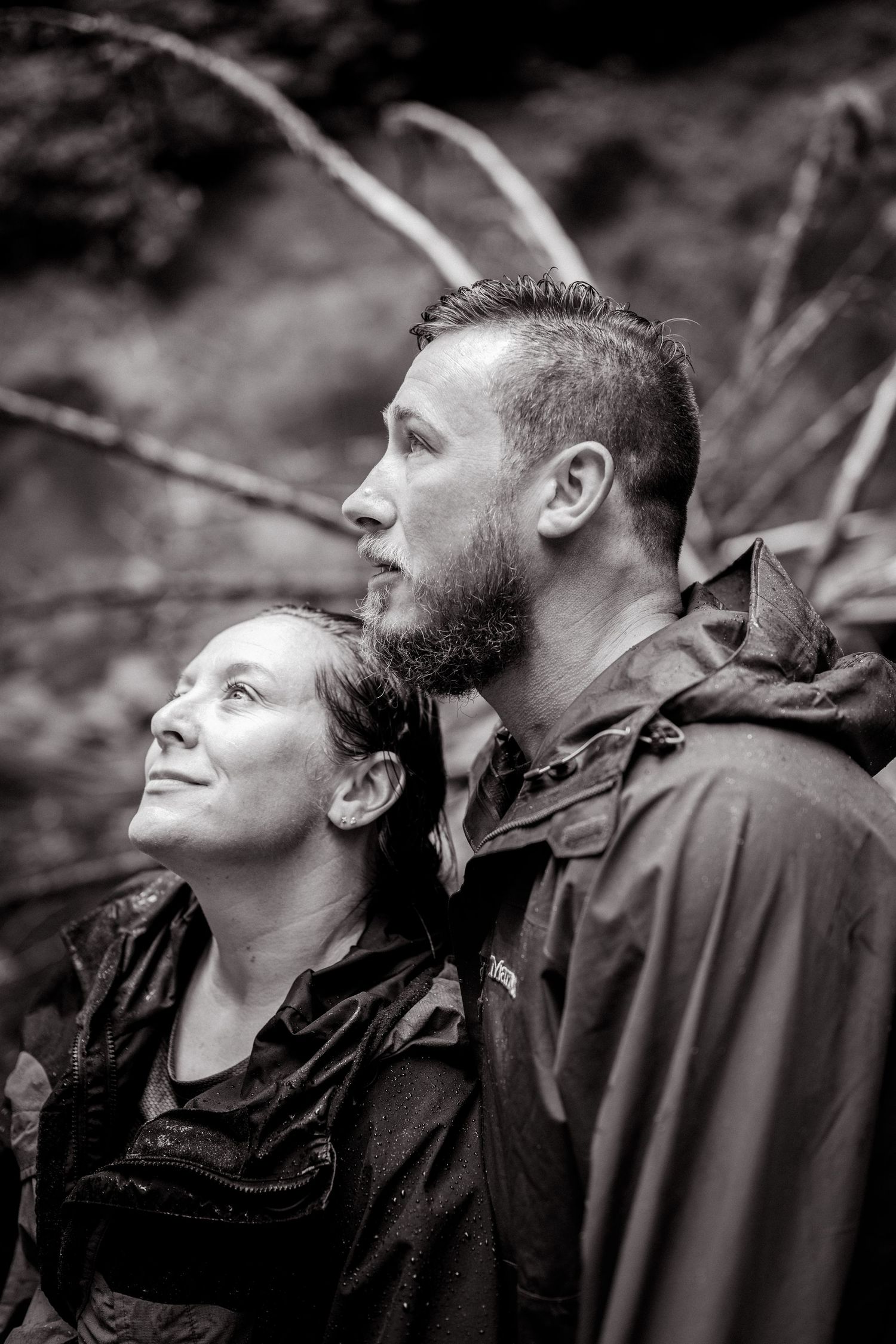 Black and white portrait of a couple in rain jackets looking up outdoors, surrounded by lush forest foliage.