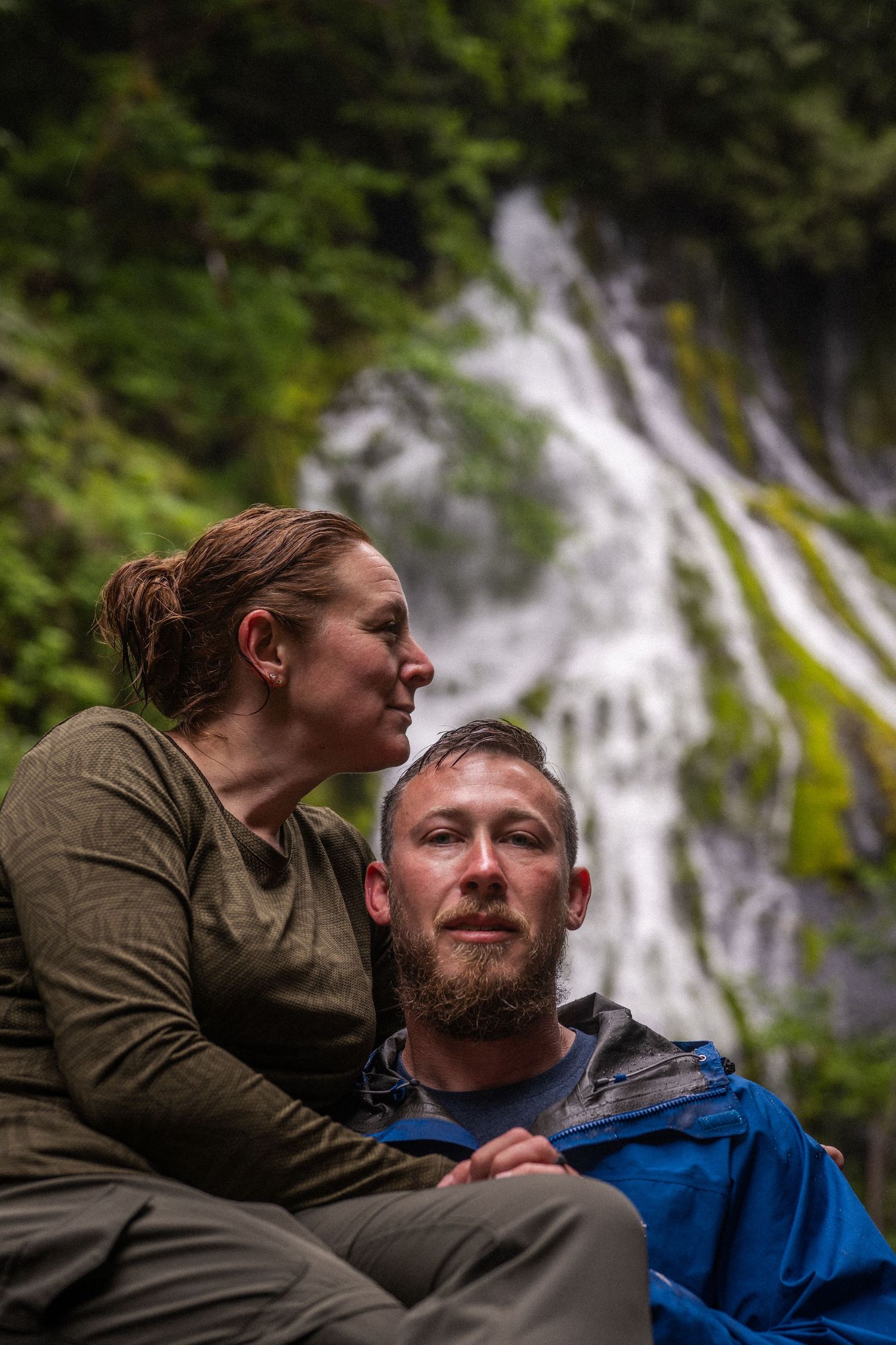 Couple sitting together in front of a cascading waterfall surrounded by lush green forest.
