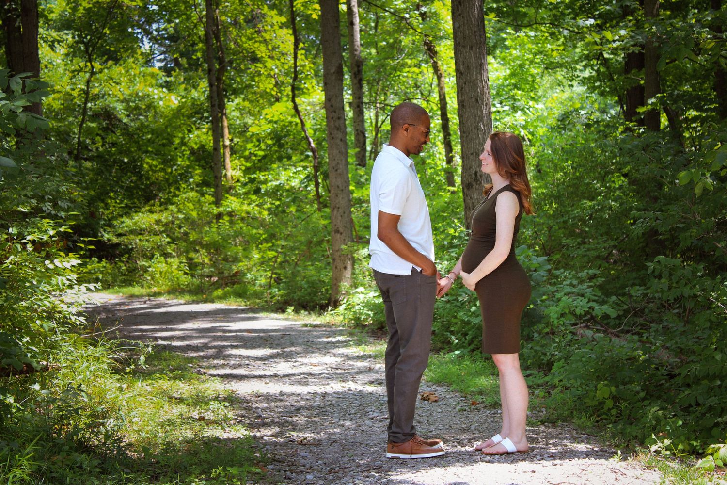 Two people stand facing each other on a sunlit gravel path surrounded by lush green forest foliage.