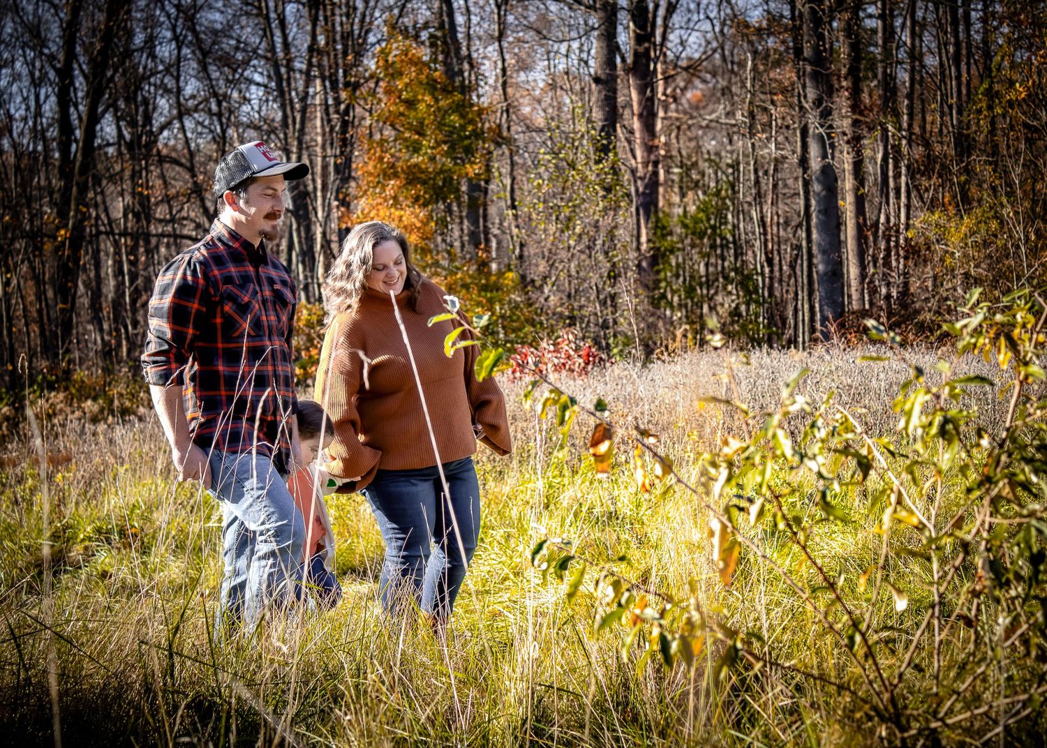 People walk through a sunlit meadow with tall grass and autumn trees in the background during golden hour.