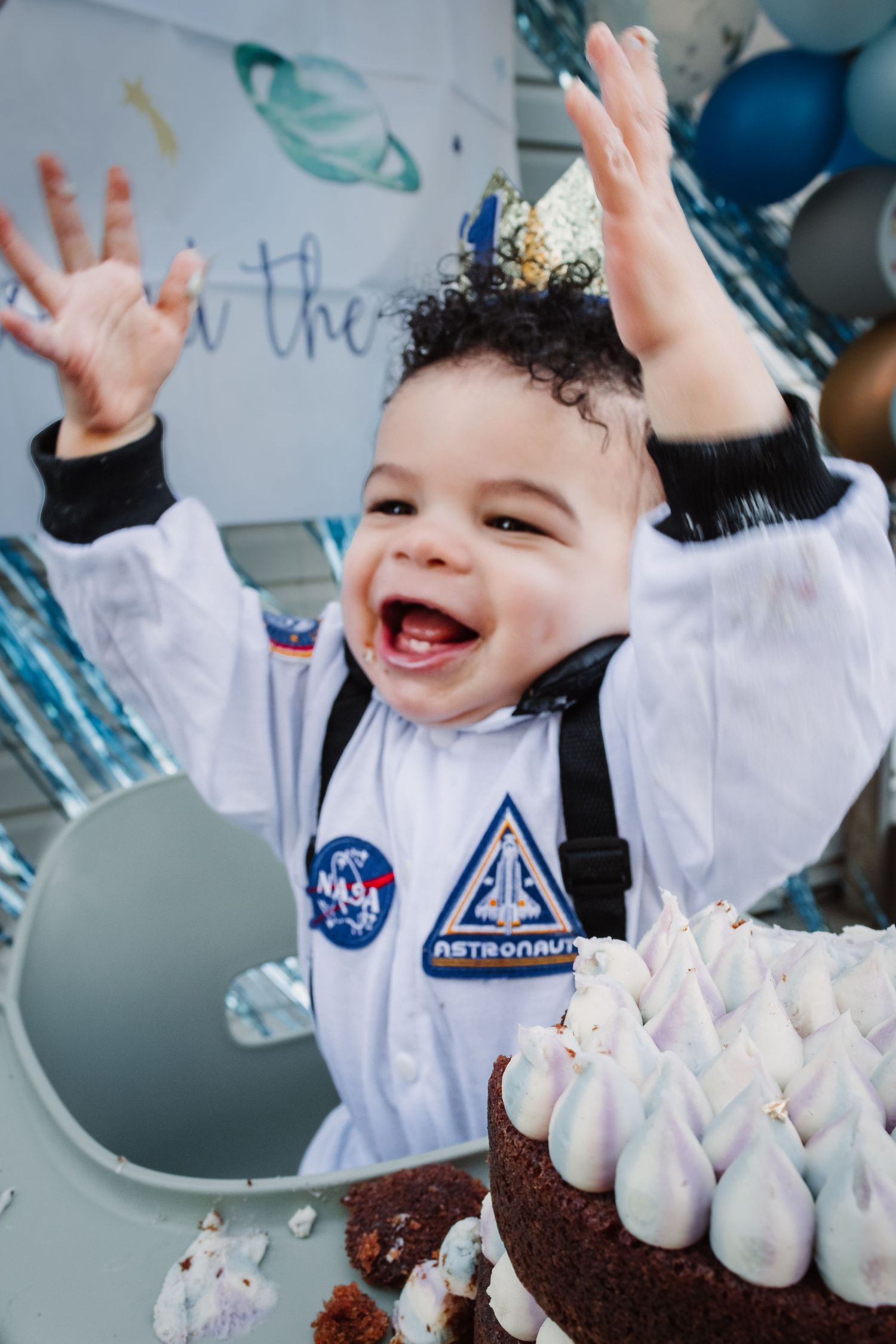 Small child in a NASA astronaut costume raises hands excitedly by a birthday cake with white frosting decorations.