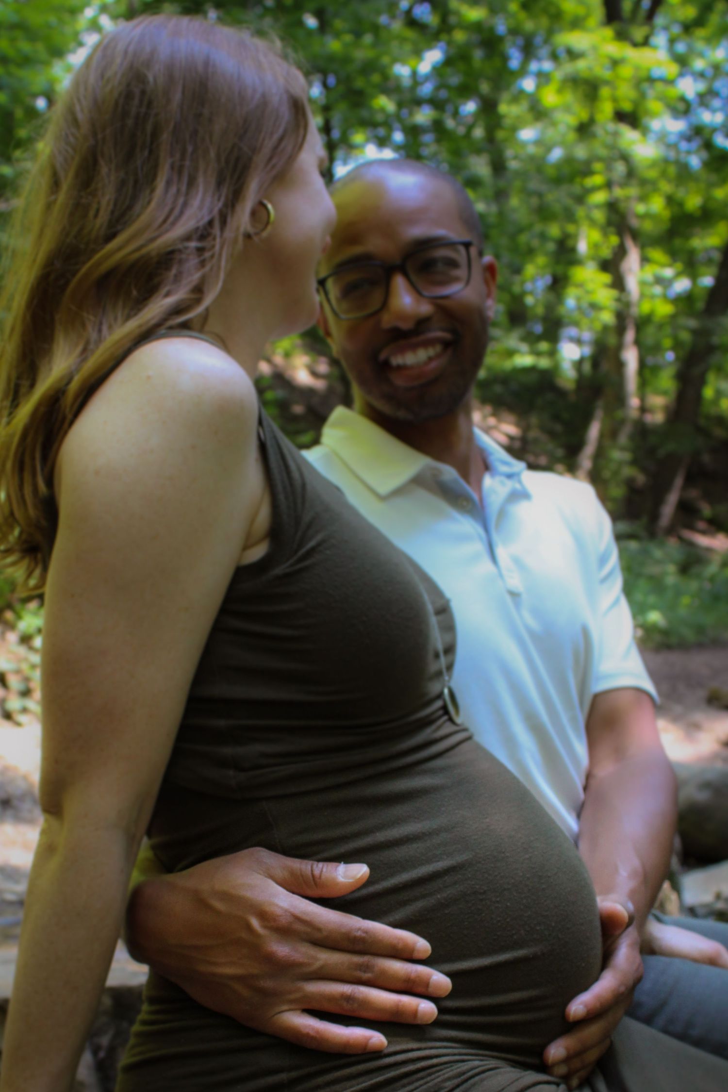 Happy couple sits together outdoors in a natural setting while sharing a tender moment in the sunshine.