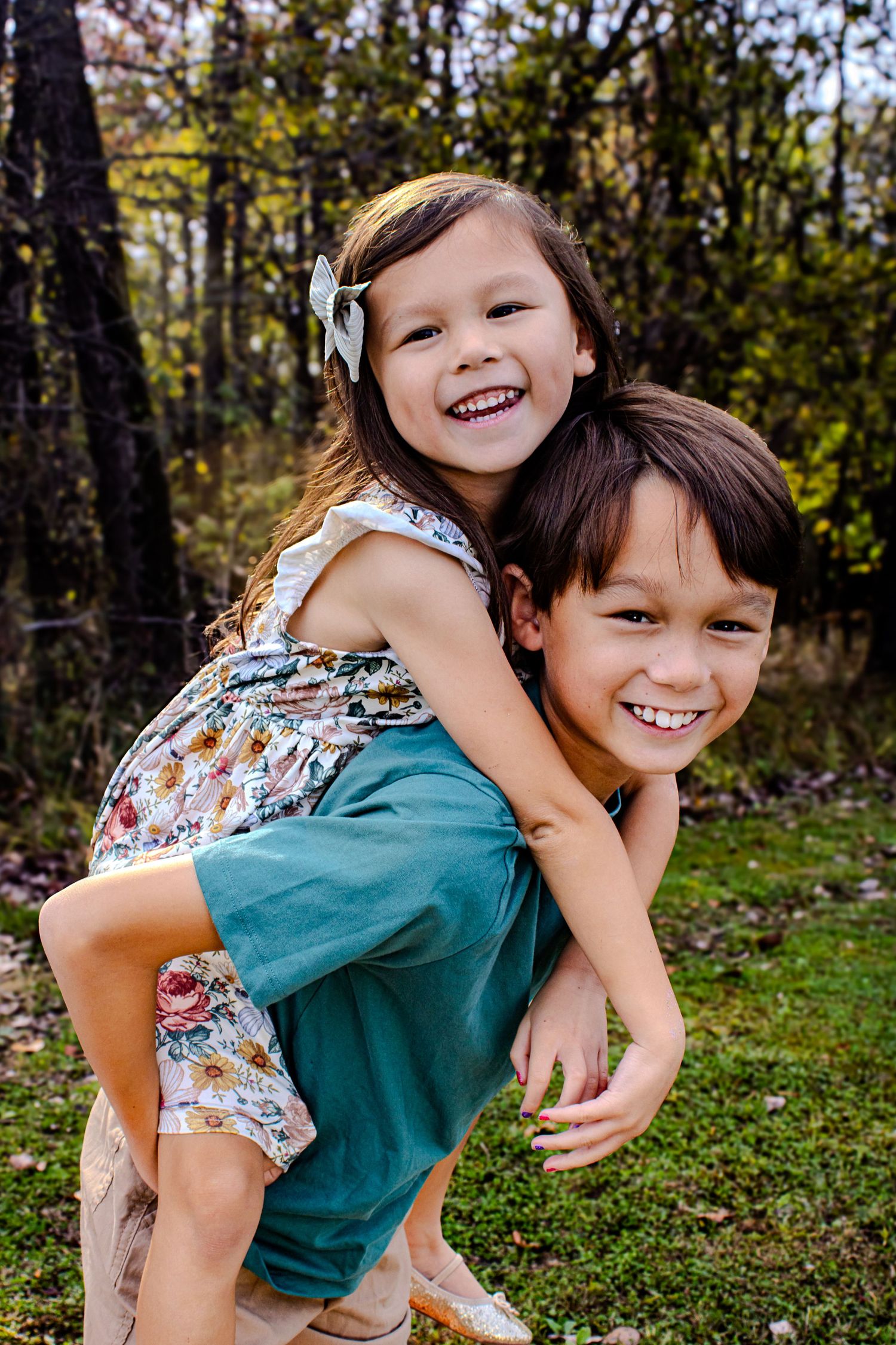 Two siblings play and laugh together in a wooded outdoor setting on a sunny day.