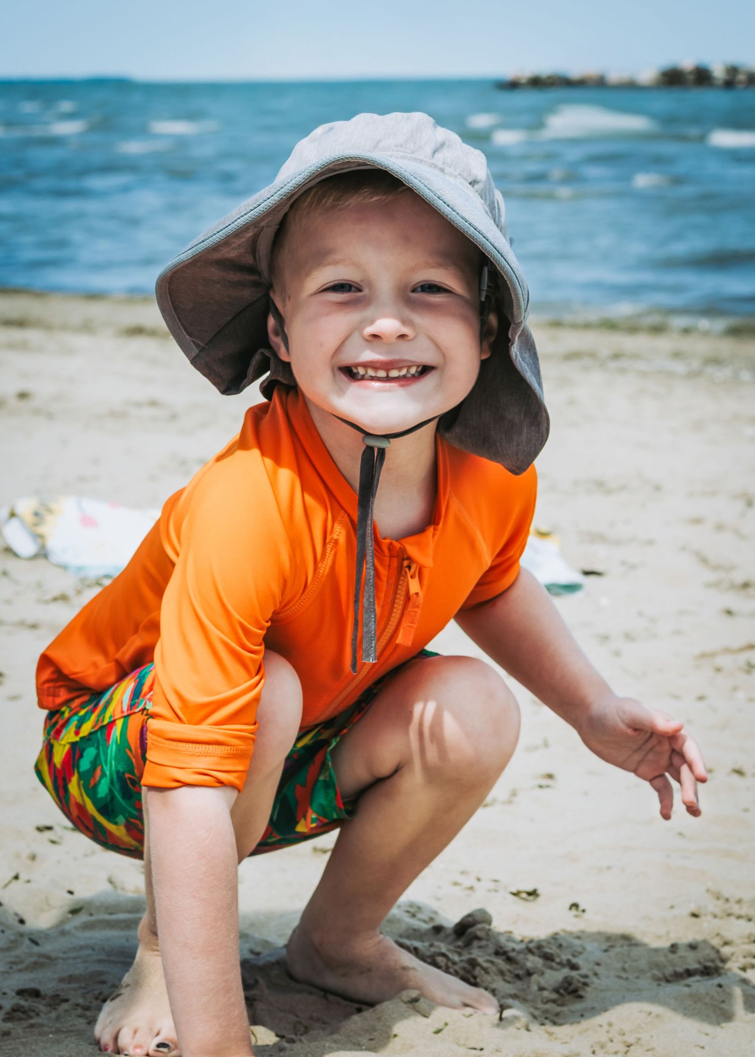 Child crouching on beach sand wearing orange swim shirt and protective hat.