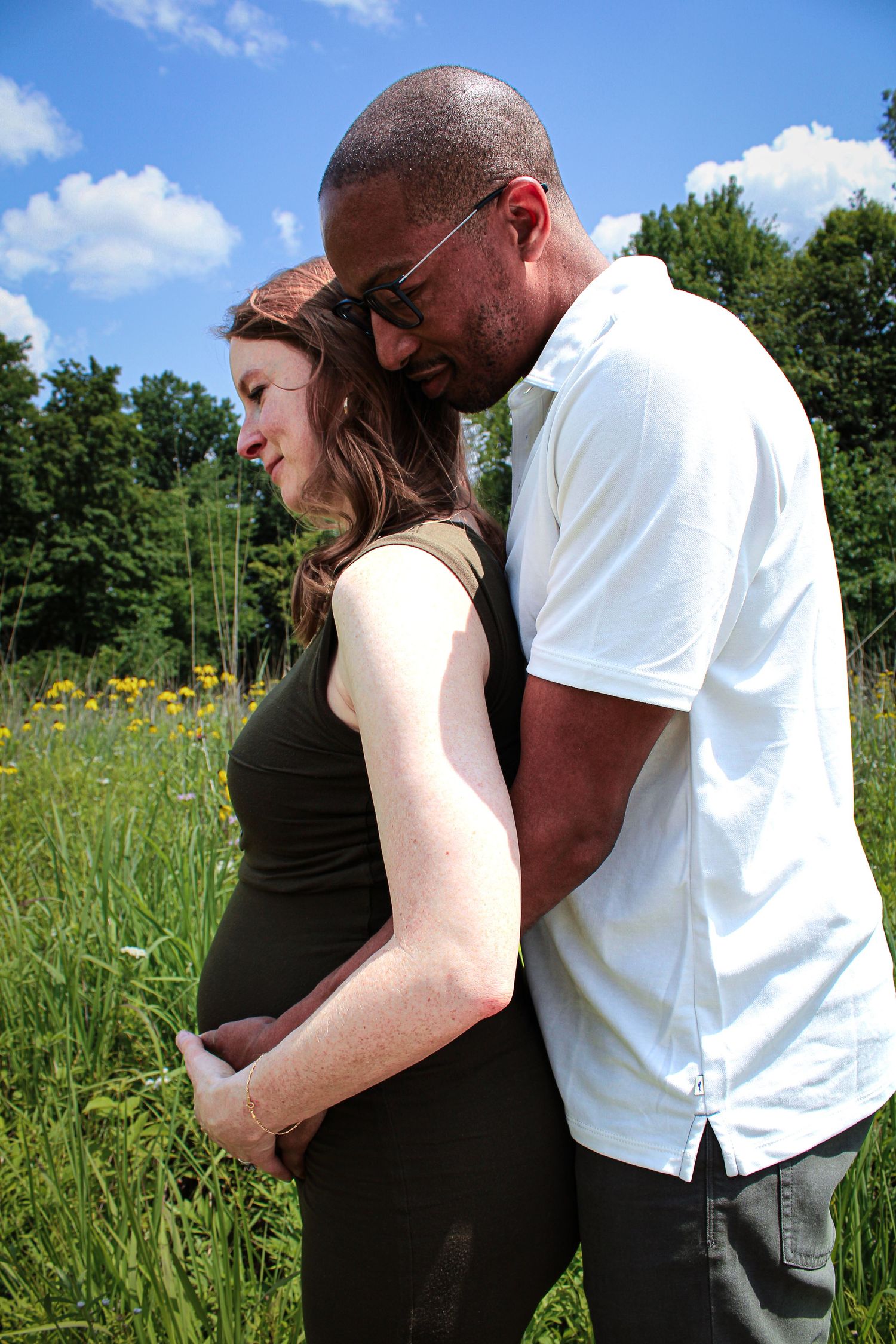 A couple shares an intimate moment in a sunny meadow with wildflowers and trees in the background.