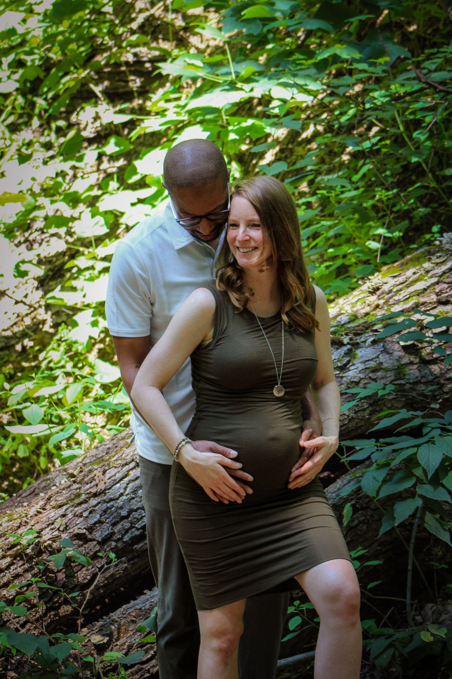 A couple shares an intimate moment during an outdoor maternity photoshoot surrounded by lush green foliage.