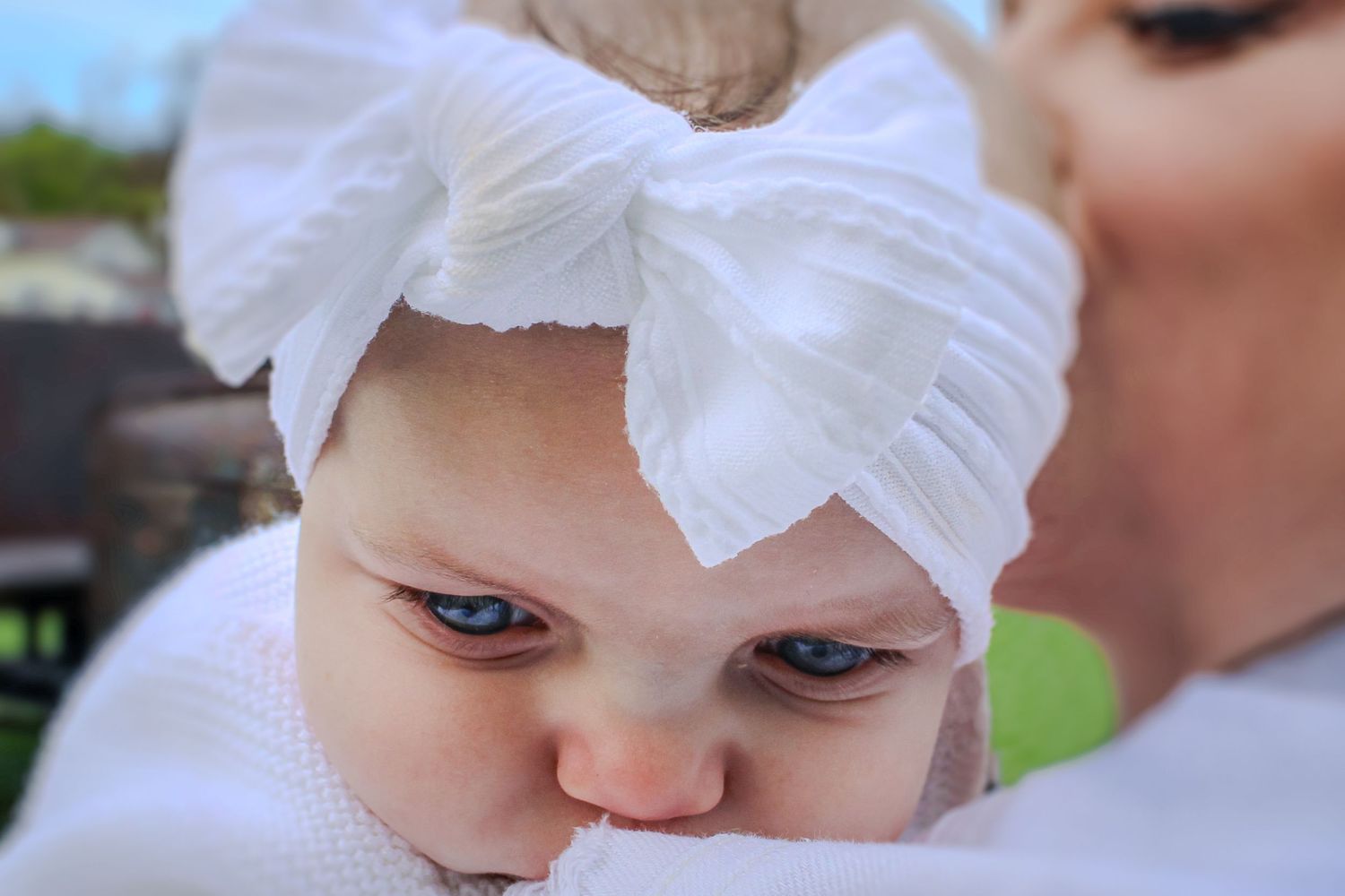 A baby wearing a large white bow headband looks at the camera with an expressive face.