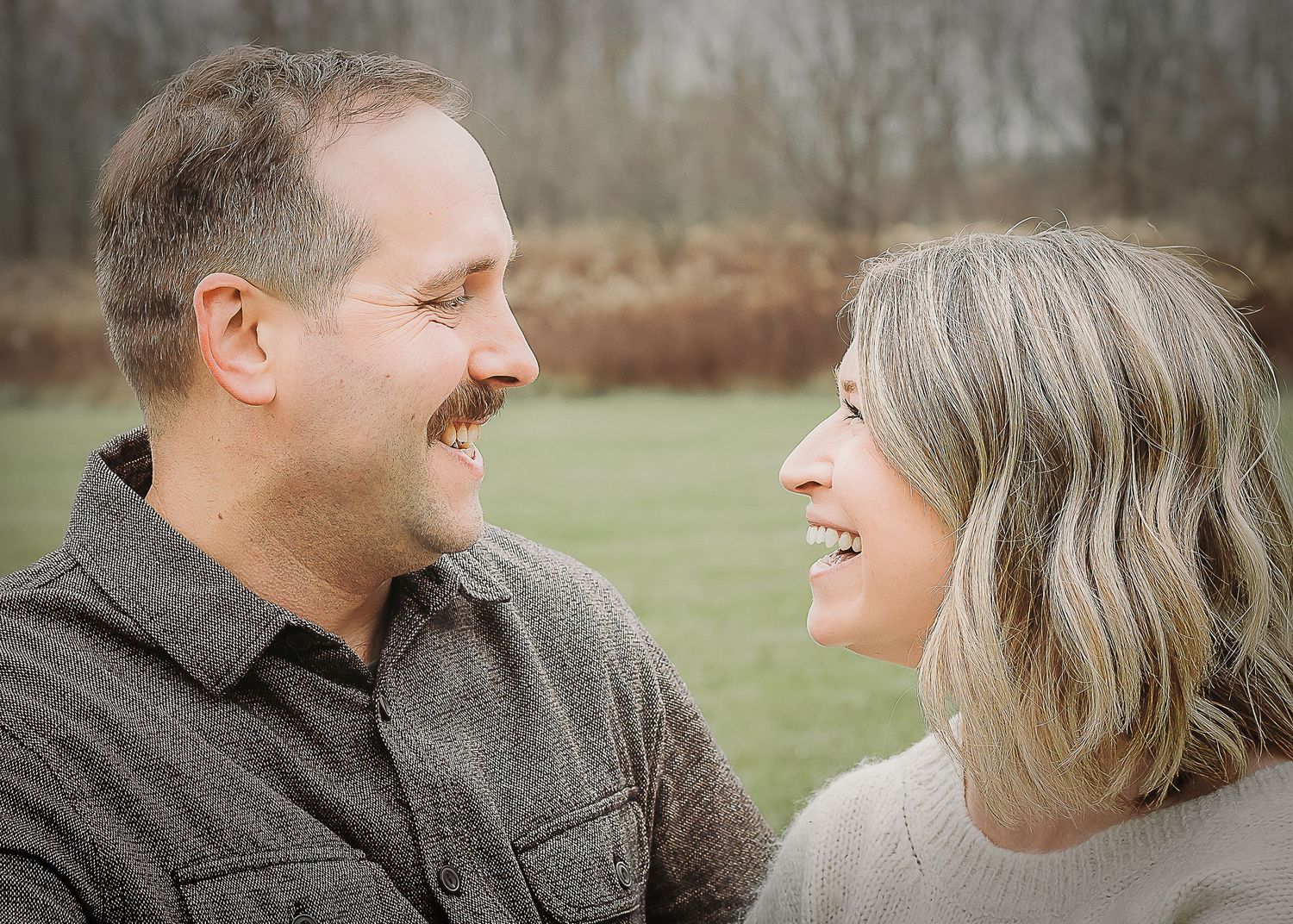 A couple laughs and smiles at each other while posing outdoors in a grassy field.