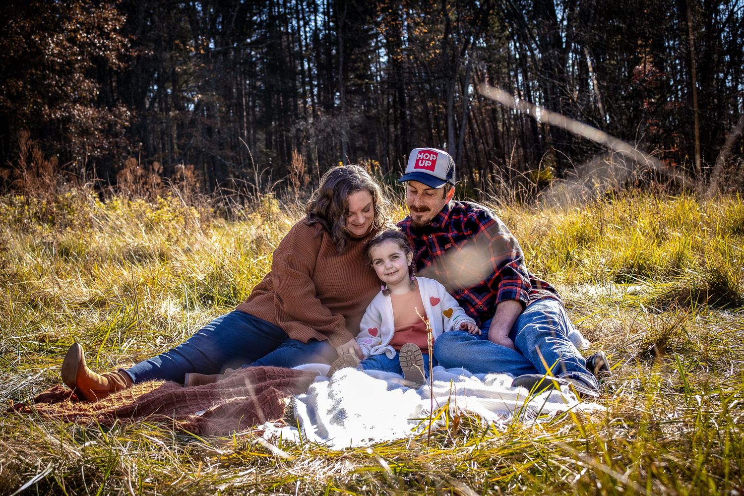 People relax and enjoy a sunny day laying on blankets in a grassy meadow surrounded by trees during fall.
