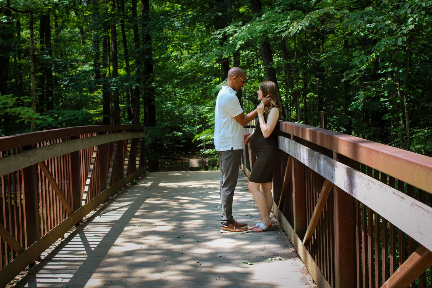 Couple sharing a romantic moment on a wooden bridge surrounded by lush green forest on a summer day.