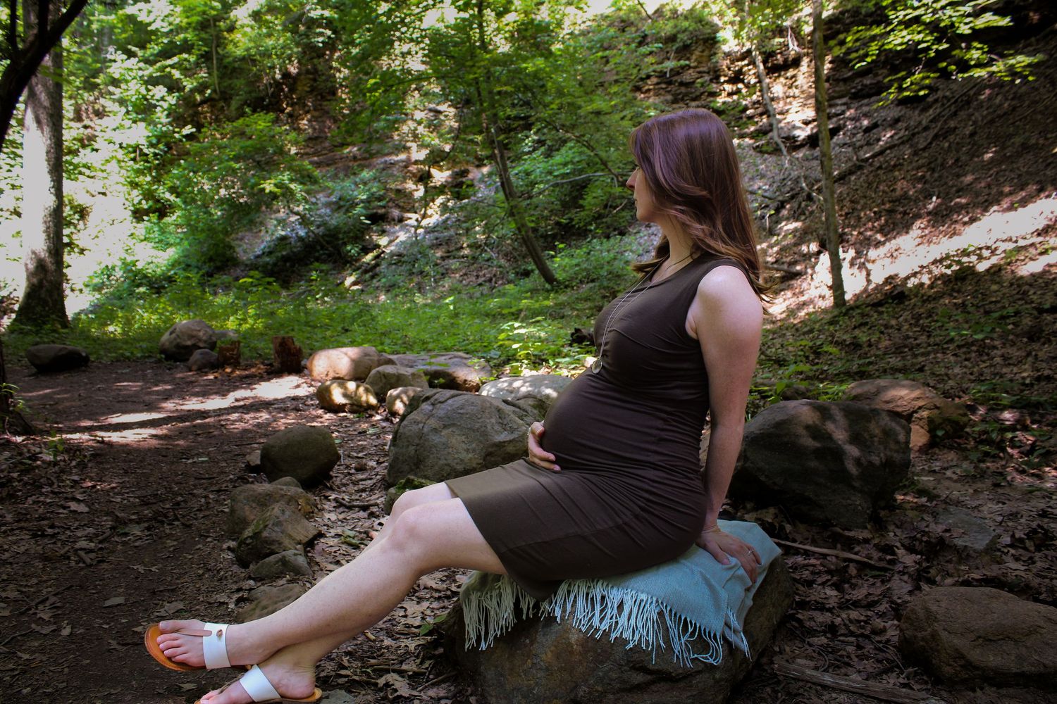 A pregnant woman in a brown dress sits on a rock in a wooded park during warm summer weather.