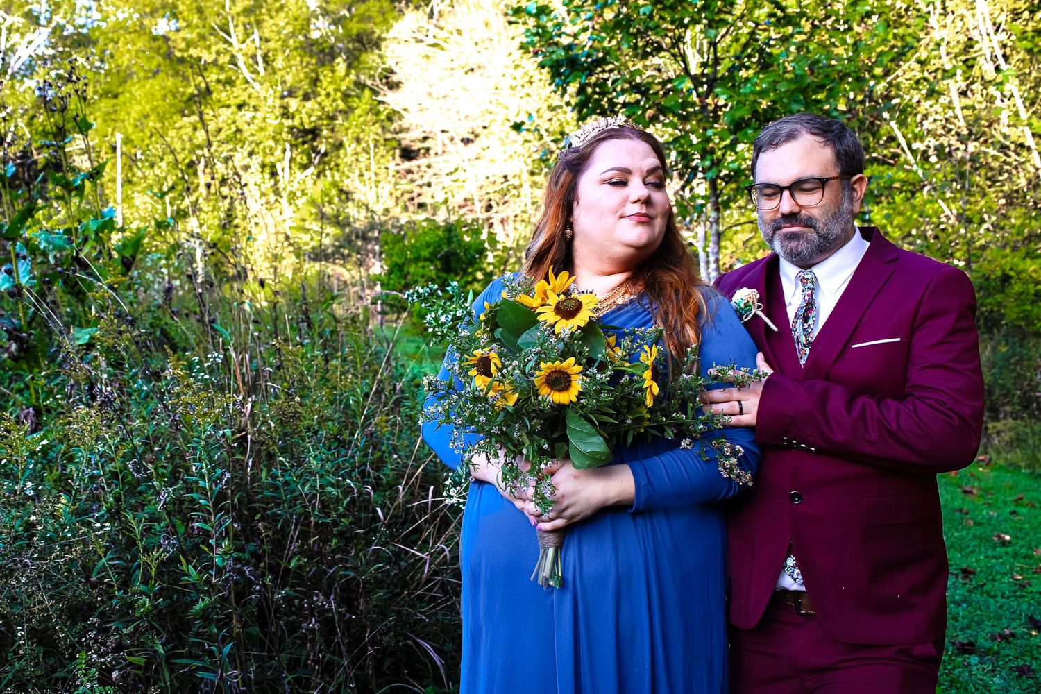 A couple wearing burgundy and blue outfits poses together in a outdoor garden setting during a romantic photoshoot.