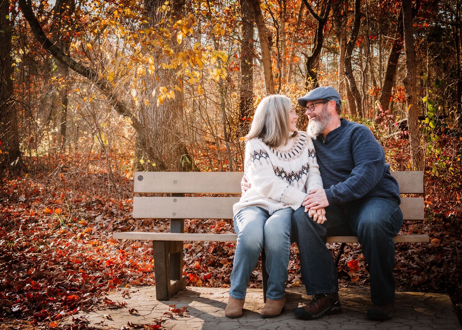 Two people share a tender moment on a wooden bench surrounded by vibrant autumn foliage in a forest setting.