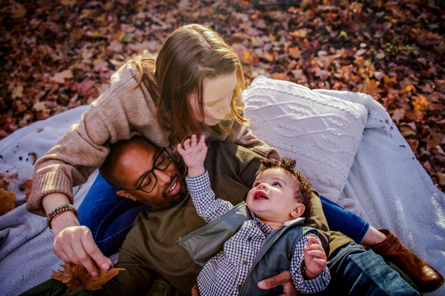 Family lies on autumn leaves in park during outdoor photo session with multiple candid moments at sunset.