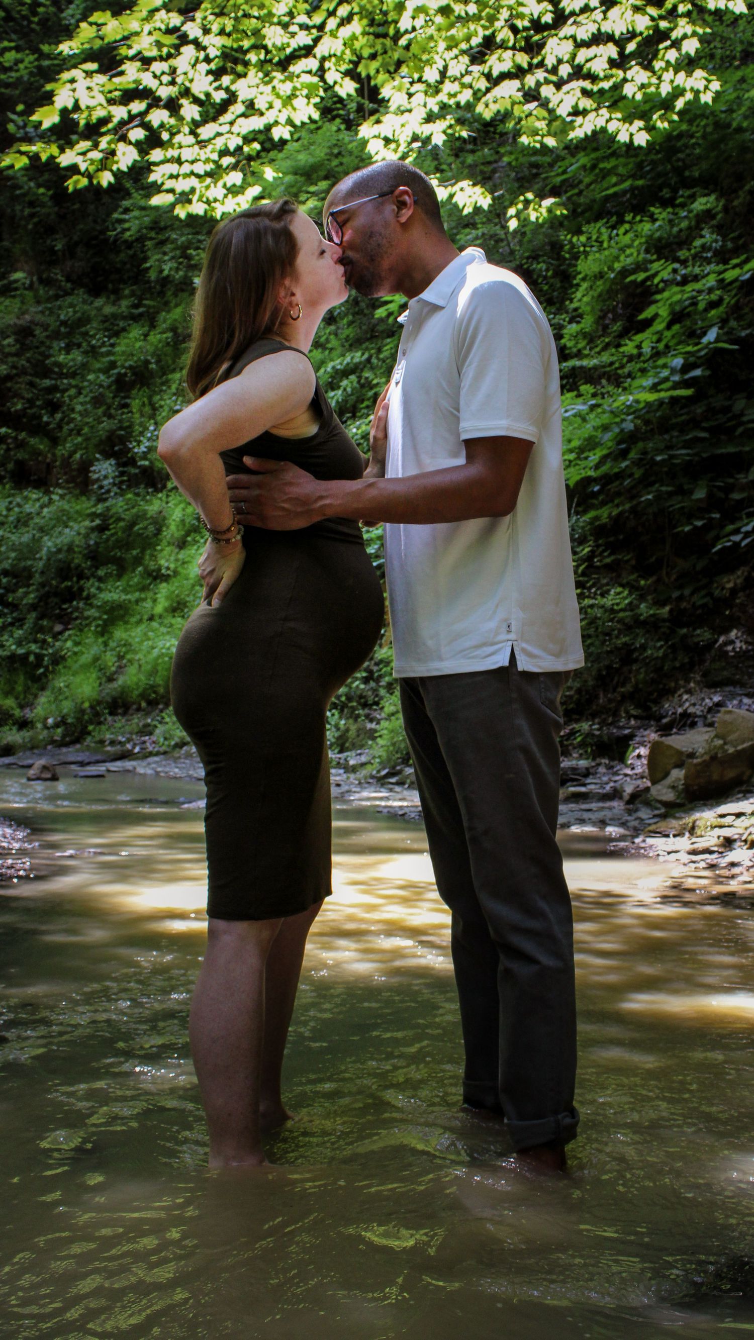 A couple shares a romantic moment while standing together in a shallow creek surrounded by lush summer foliage.